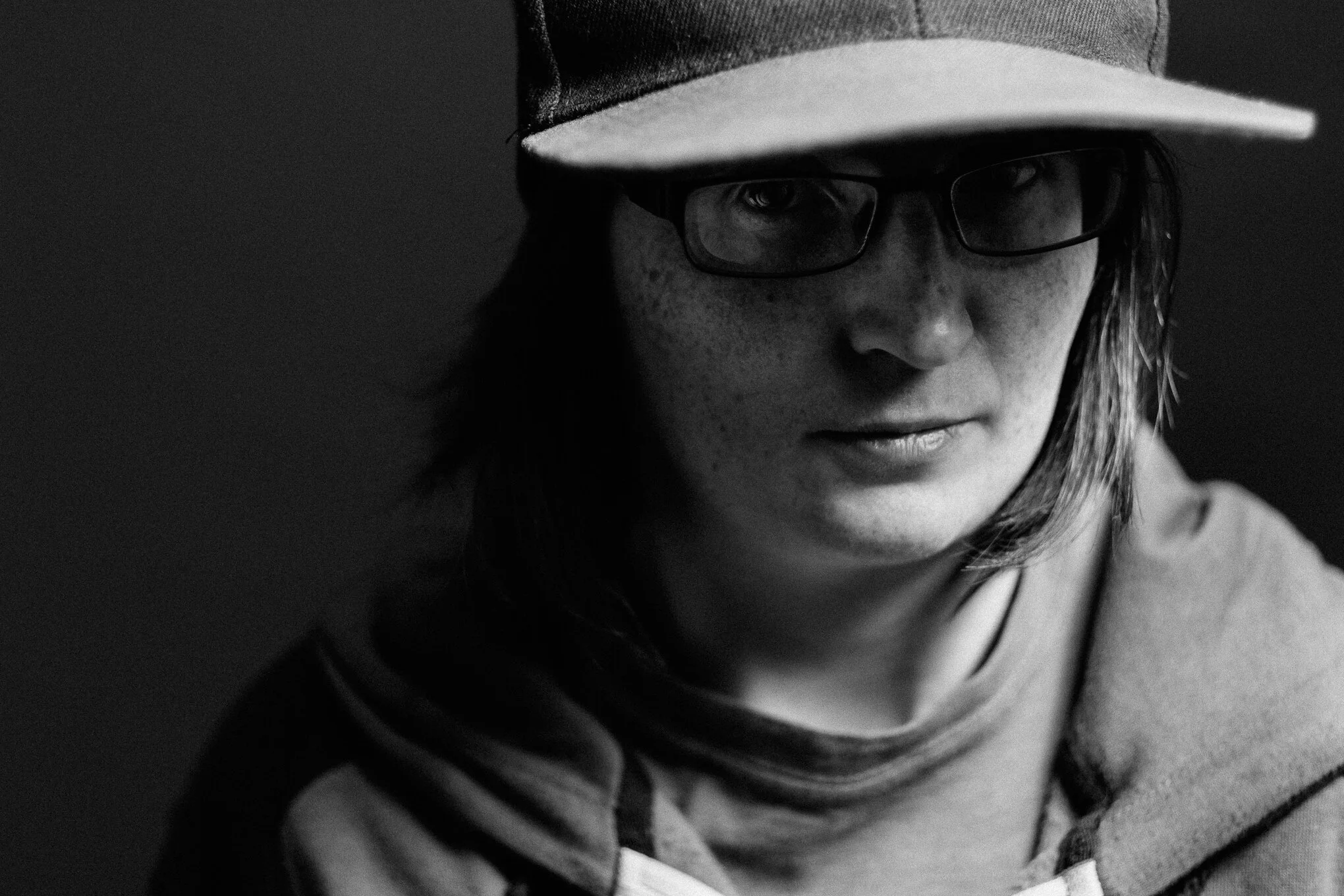 A black and white image of a woman wearing glasses and a baseball cap, looking downward with a serious expression.
