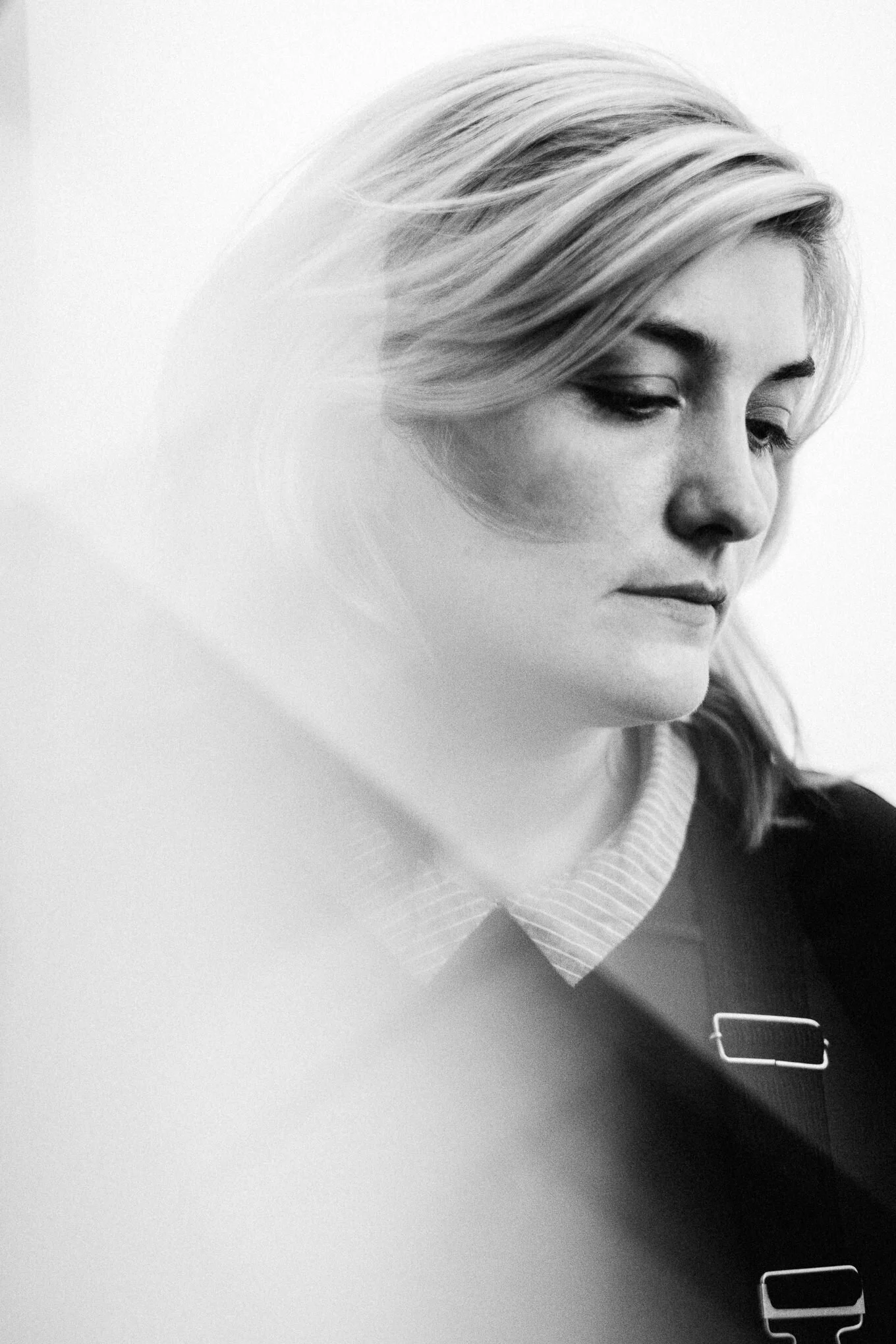 A black-and-white photograph of a woman with light-colored hair looking down, wearing a striped shirt and a dark jacket with a visible zipper.