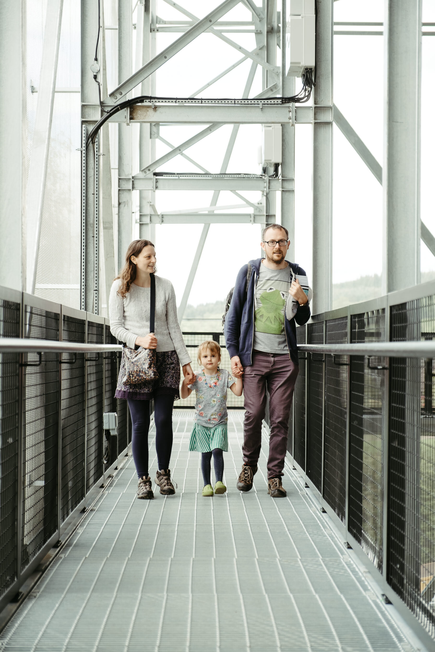 A family of four, including two children and two adults, walking together on an outdoor metal bridge. The man is wearing glasses and a striped t-shirt with a blue hoodie, holding hands with a young girl in a floral dress. The woman, dressed in a whit