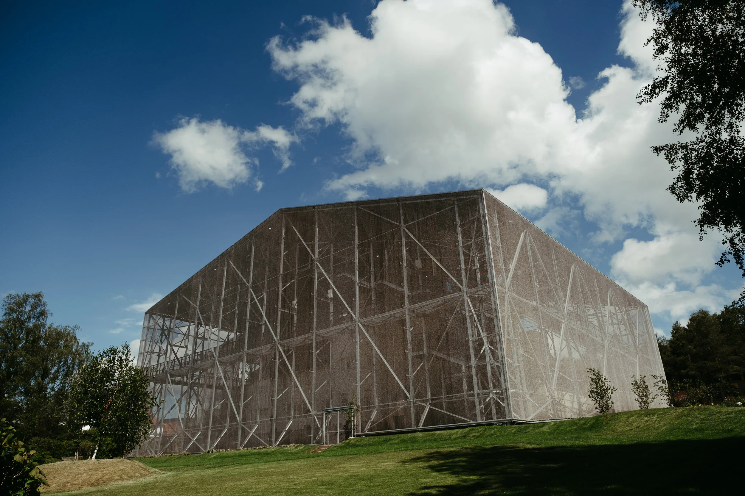 Modern building with a glass and metal exterior under a partly cloudy blue sky, surrounded by green trees and grass.
