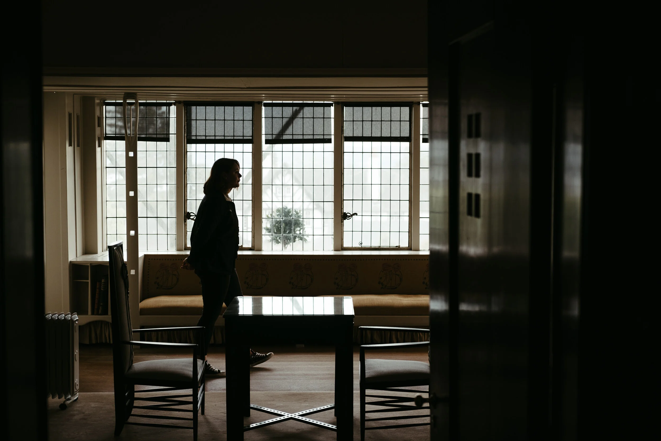 A woman standing in a dimly lit room with large grid windows behind her, with silhouettes of furniture and decor visible.