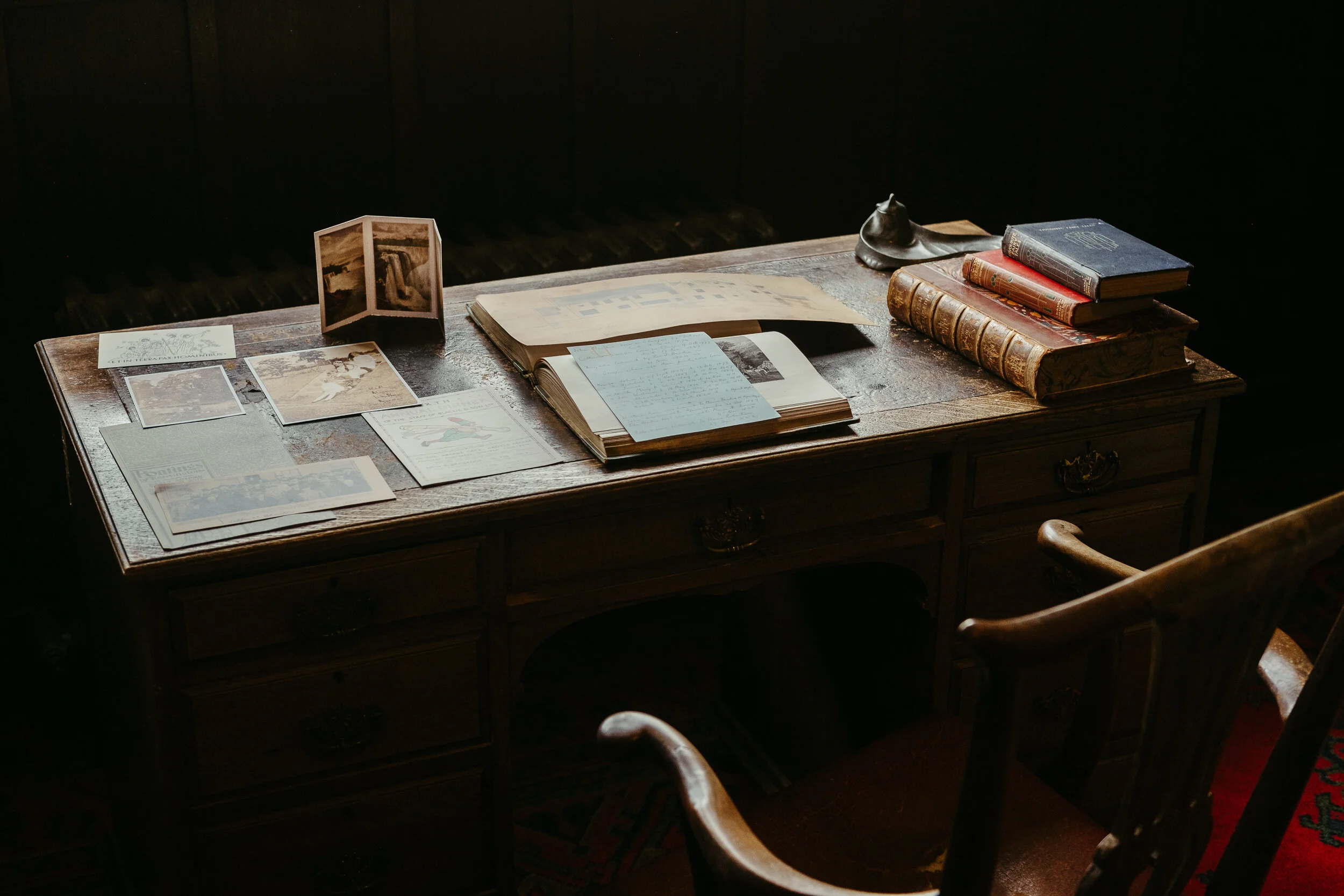 A wooden desk with vintage books, handwritten notes, photographs, and a small foldable picture frame, illuminated by soft natural light.