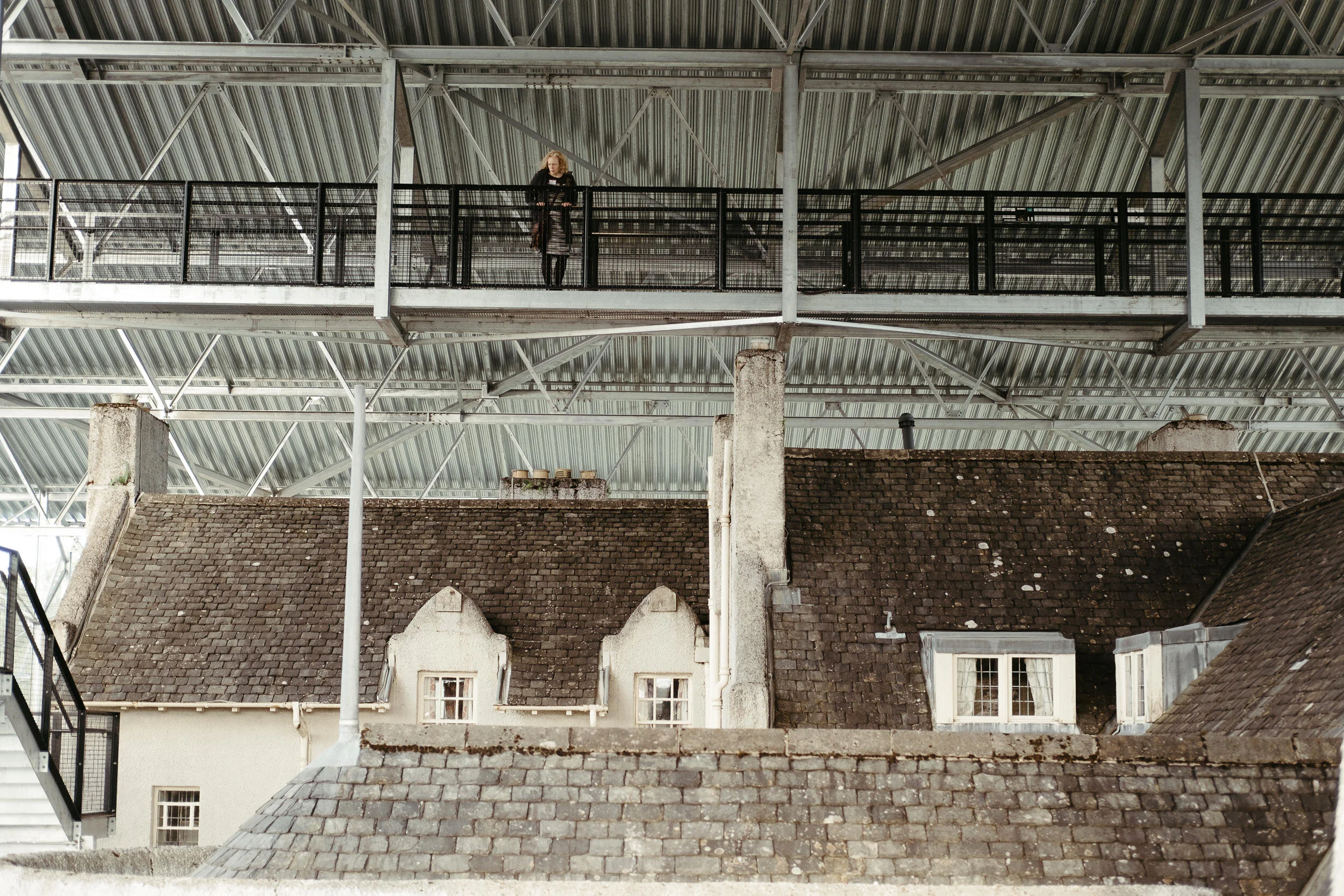 A woman standing on a modern metal and glass balcony high above an old brick building with sloped roofs in an urban setting.