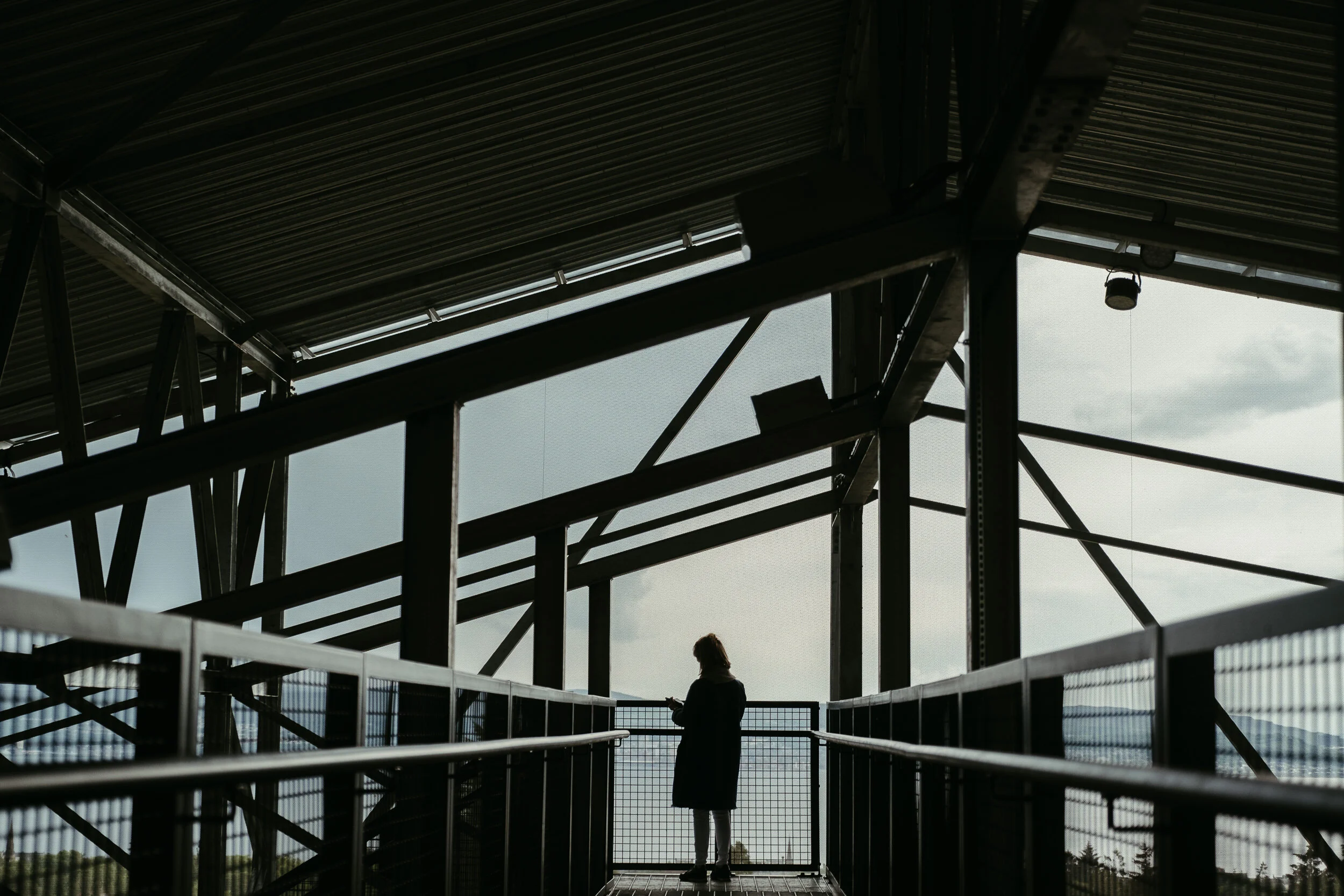 A person standing on a metal walkway under a large, partially open metal roof, with mountains and cloudy sky in the background.