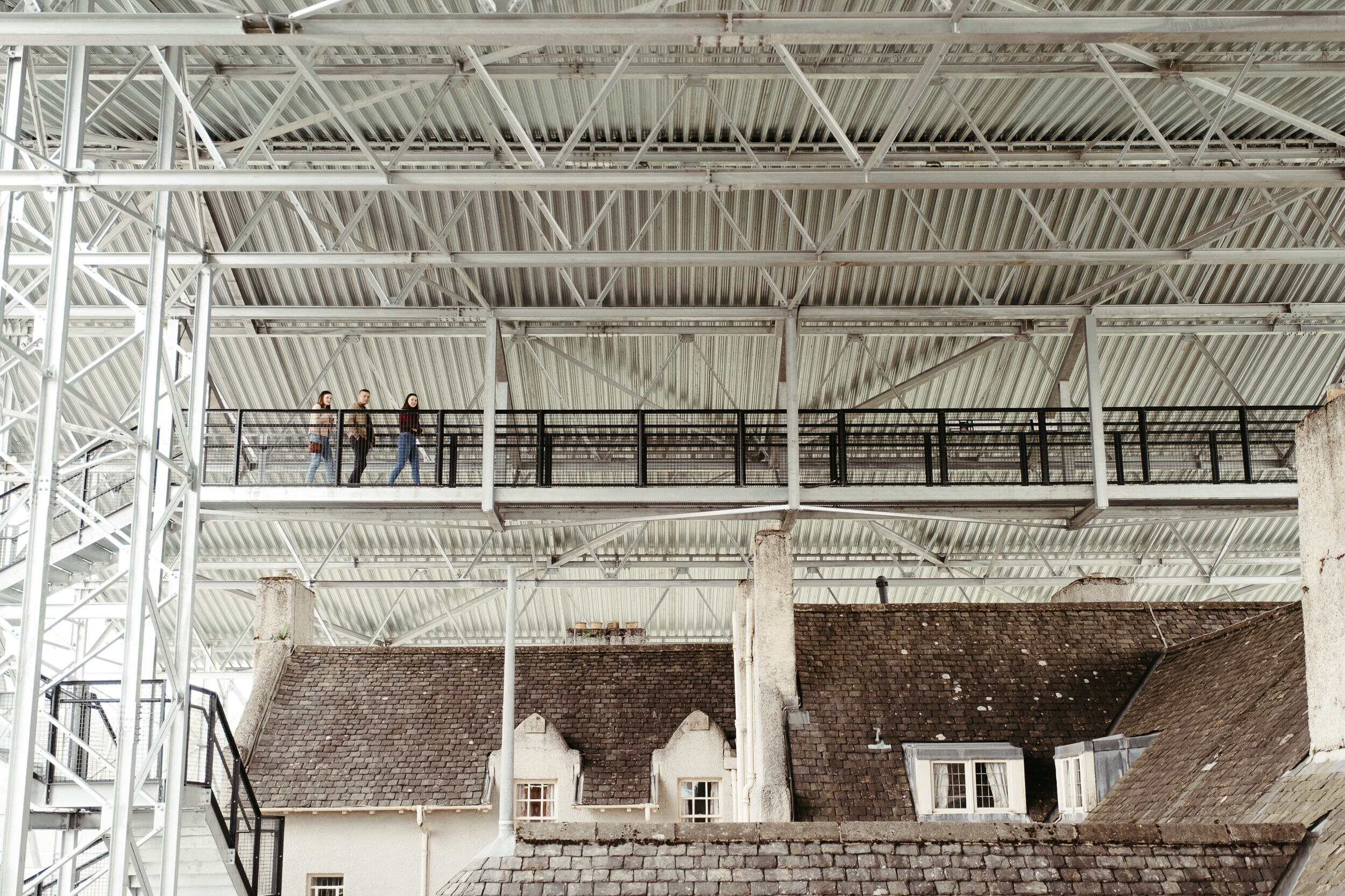 Three people walking along a metal balcony above old buildings with tiled roofs.