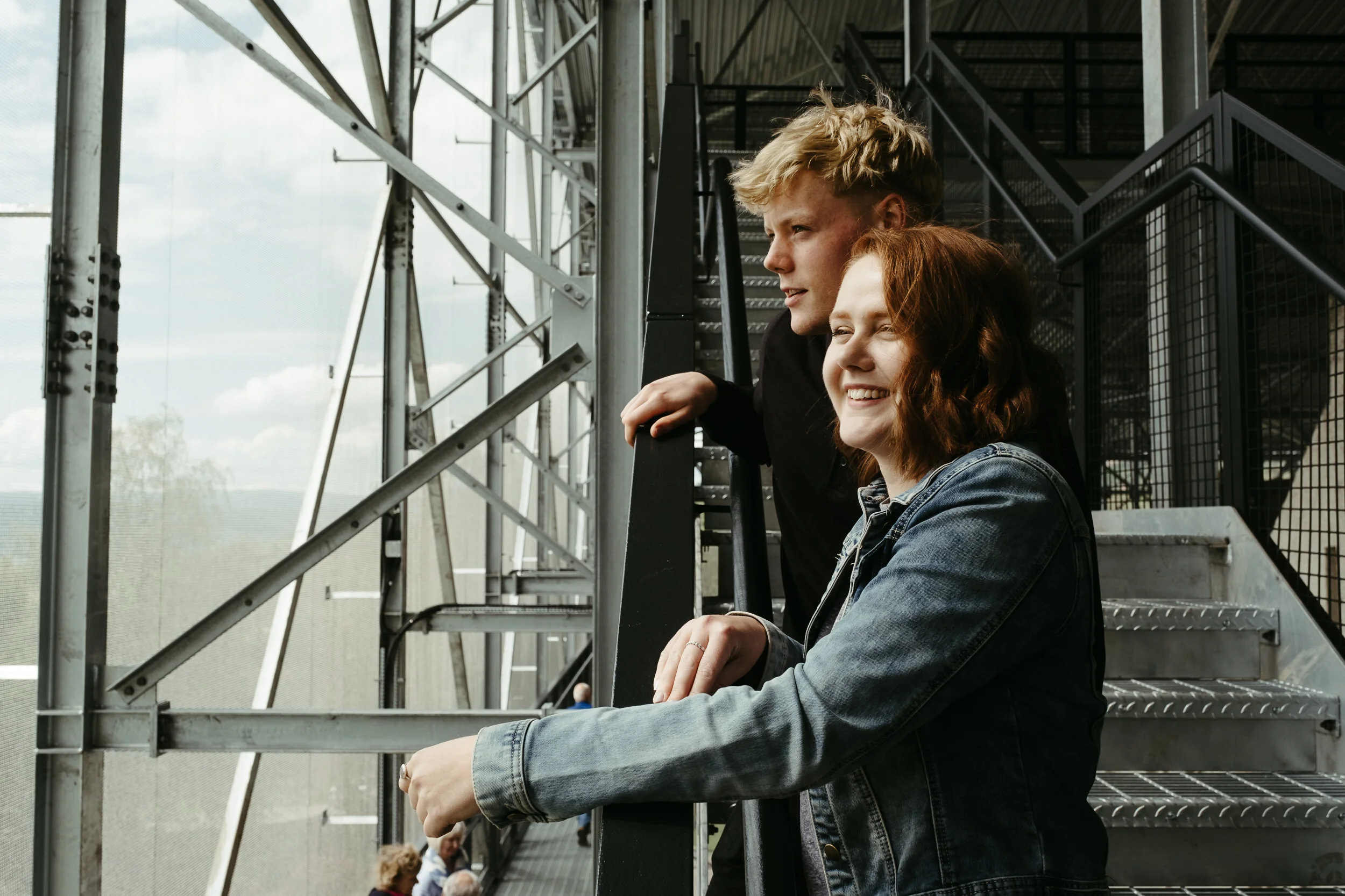 Two young women with curly hair, one with blonde and the other with red hair, sitting on metal stairs inside an industrial-looking structure, looking outside.{"type":"object","properties":{"imageAltText":"Two young women with curly hair, one with blo