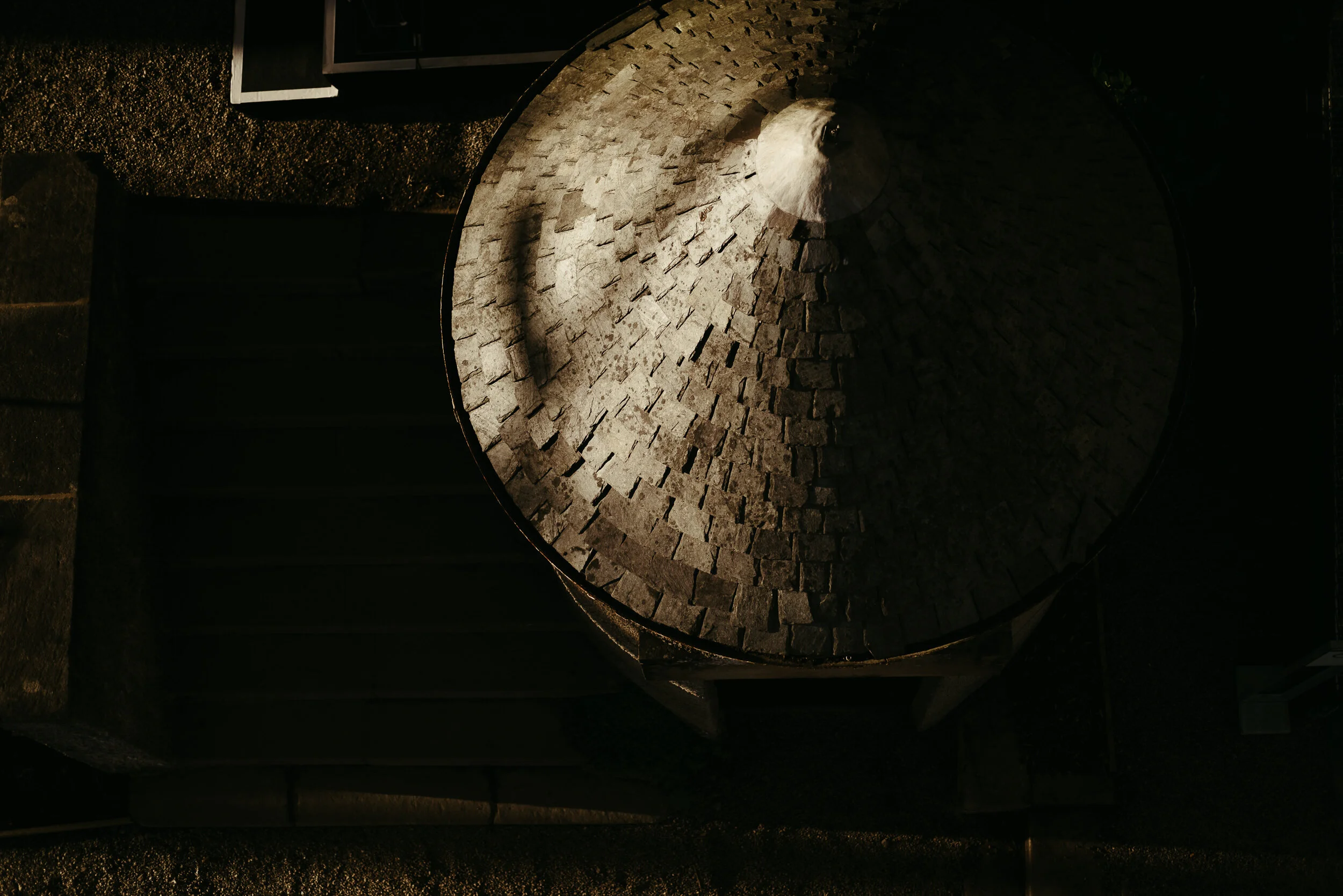 A circular stone structure with an opening in the center, appearing to be part of an outdoor fire pit or oven, surrounded by gravel on the ground, with shadowed neighboring objects.