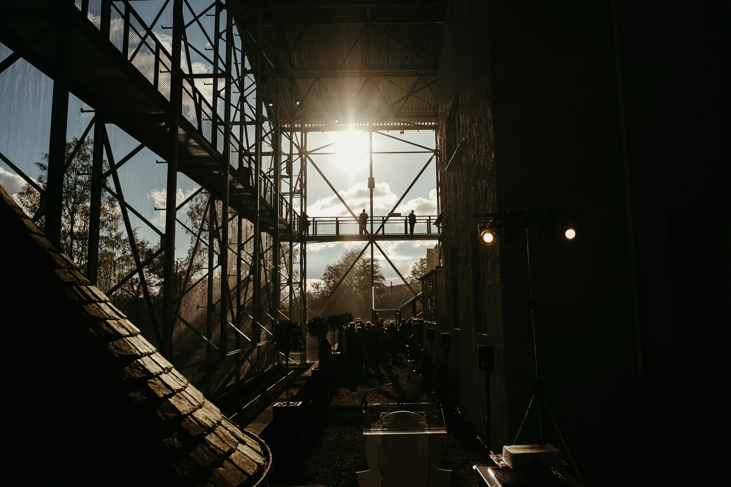 People standing on a walkway with a metal railing, silhouetted against the bright sun shining through the scaffolding of a building structure during sunset or sunrise.