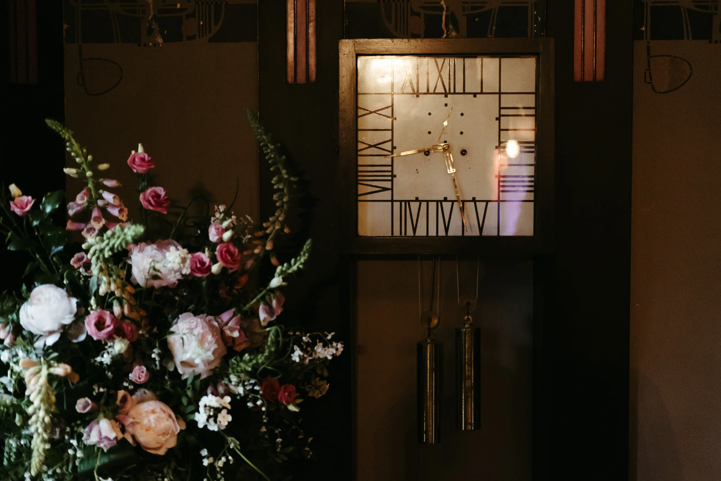 A vintage clock with Roman numerals and gold hands on a wall, with a bouquet of pink and white flowers in the foreground.