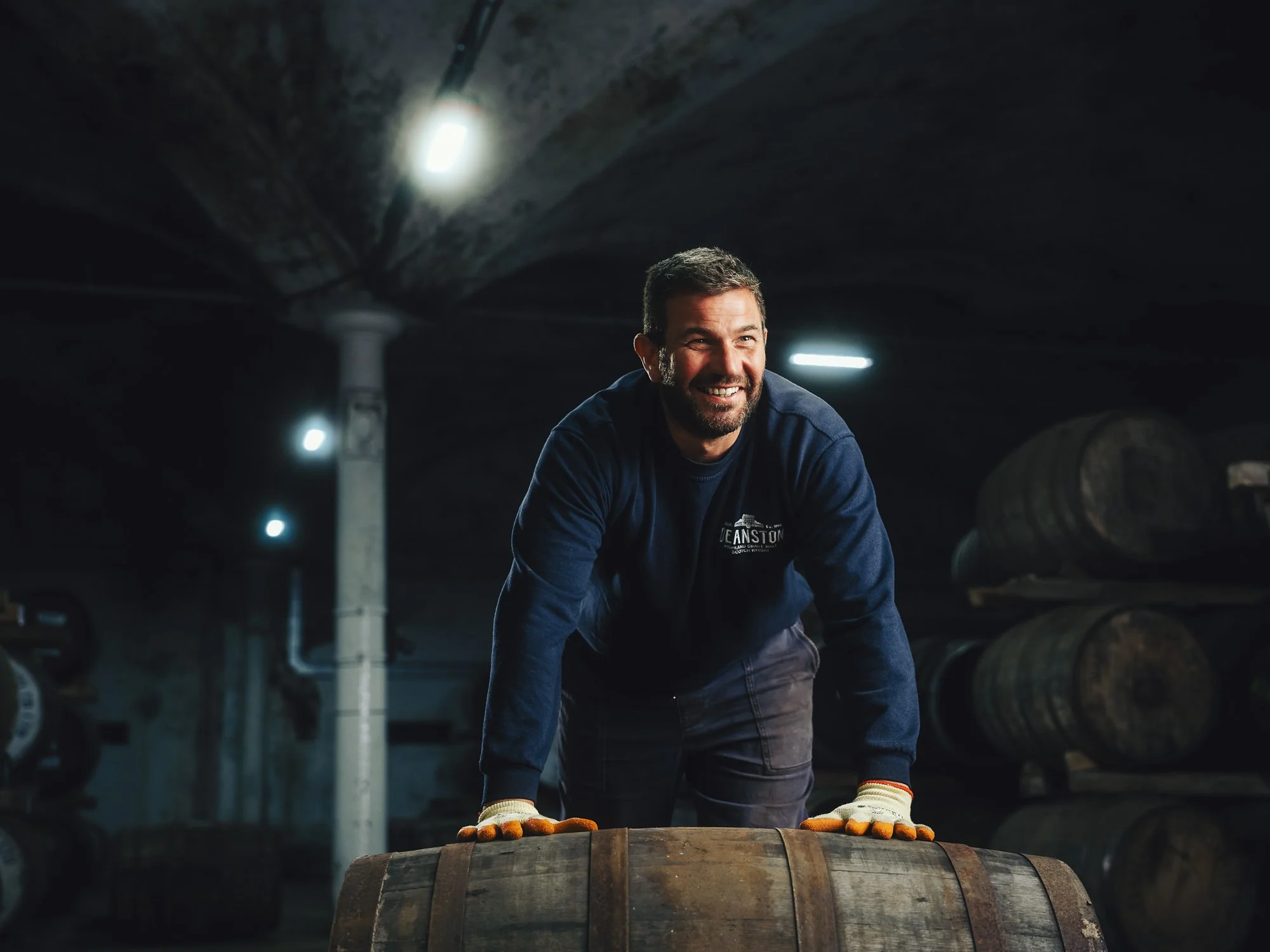 A man with a beard and dark hair smiling while working in a dimly lit cellar, wearing gloves and a dark blue shirt with a logo, standing behind a wooden barrel.