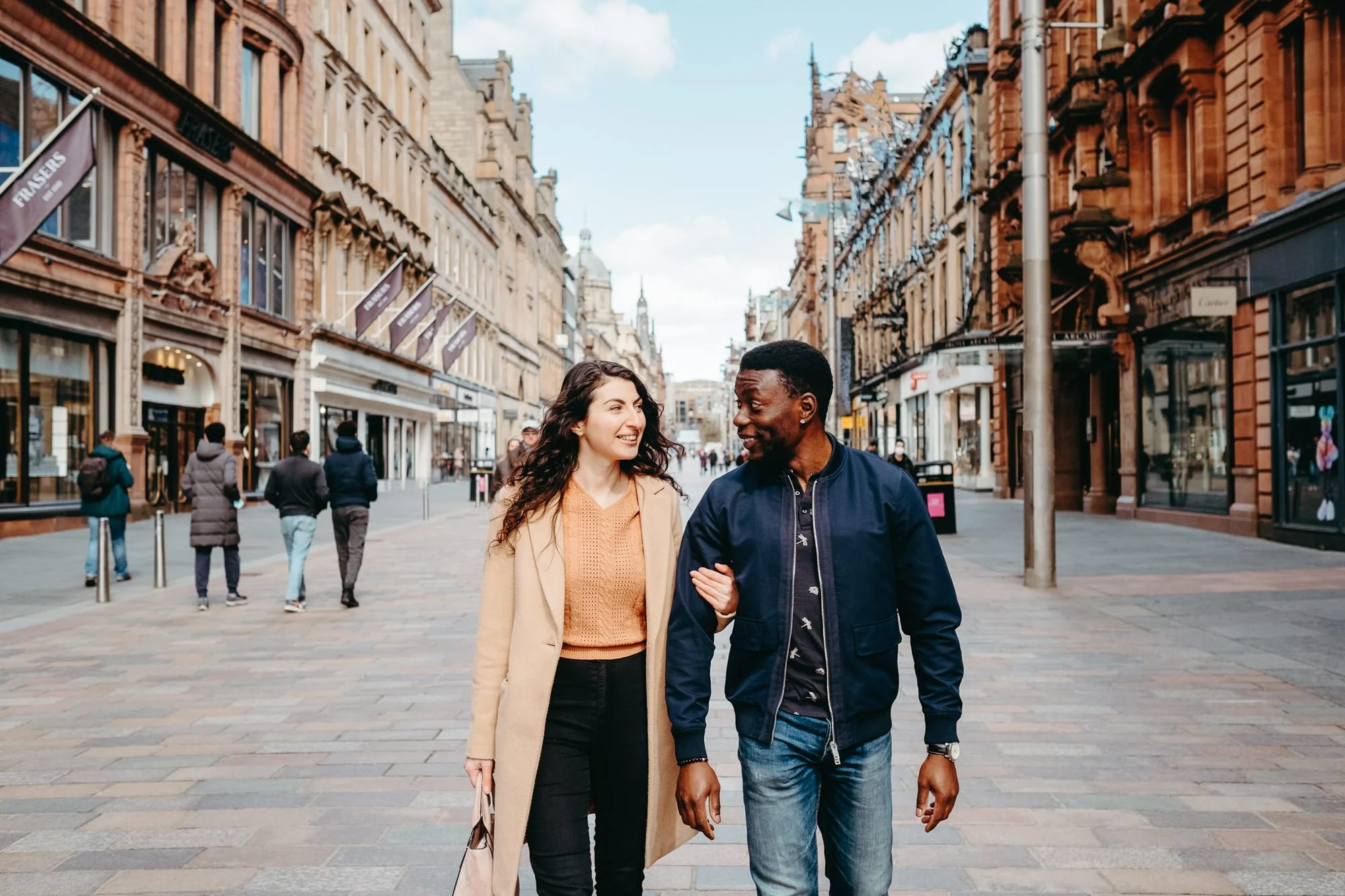 A woman and a man walking arm-in-arm down a busy shopping street, smiling and talking to each other, with people walking behind them and historic buildings lining the street.