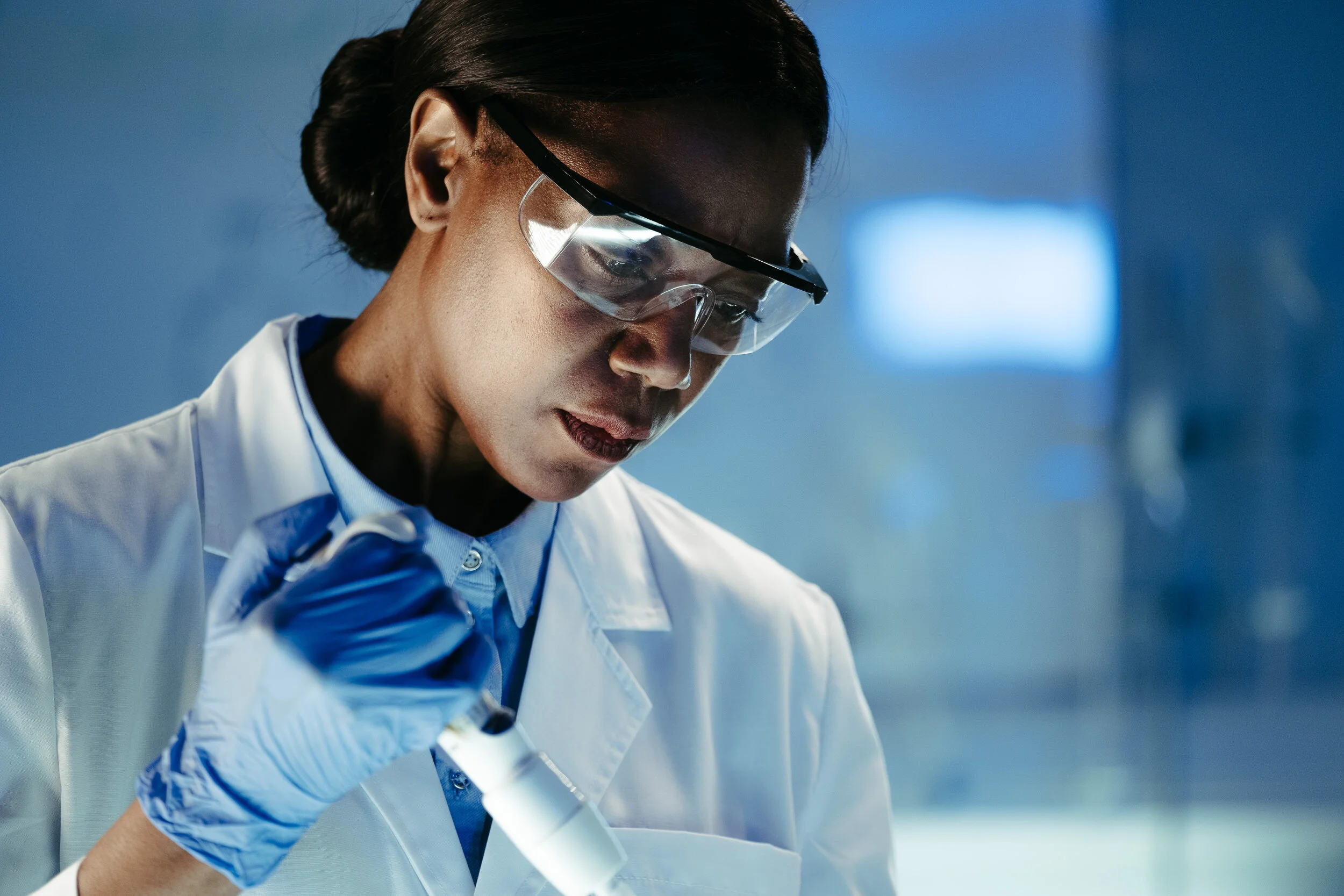 A scientist wearing safety goggles and a white lab coat looks through a microscope in a laboratory.