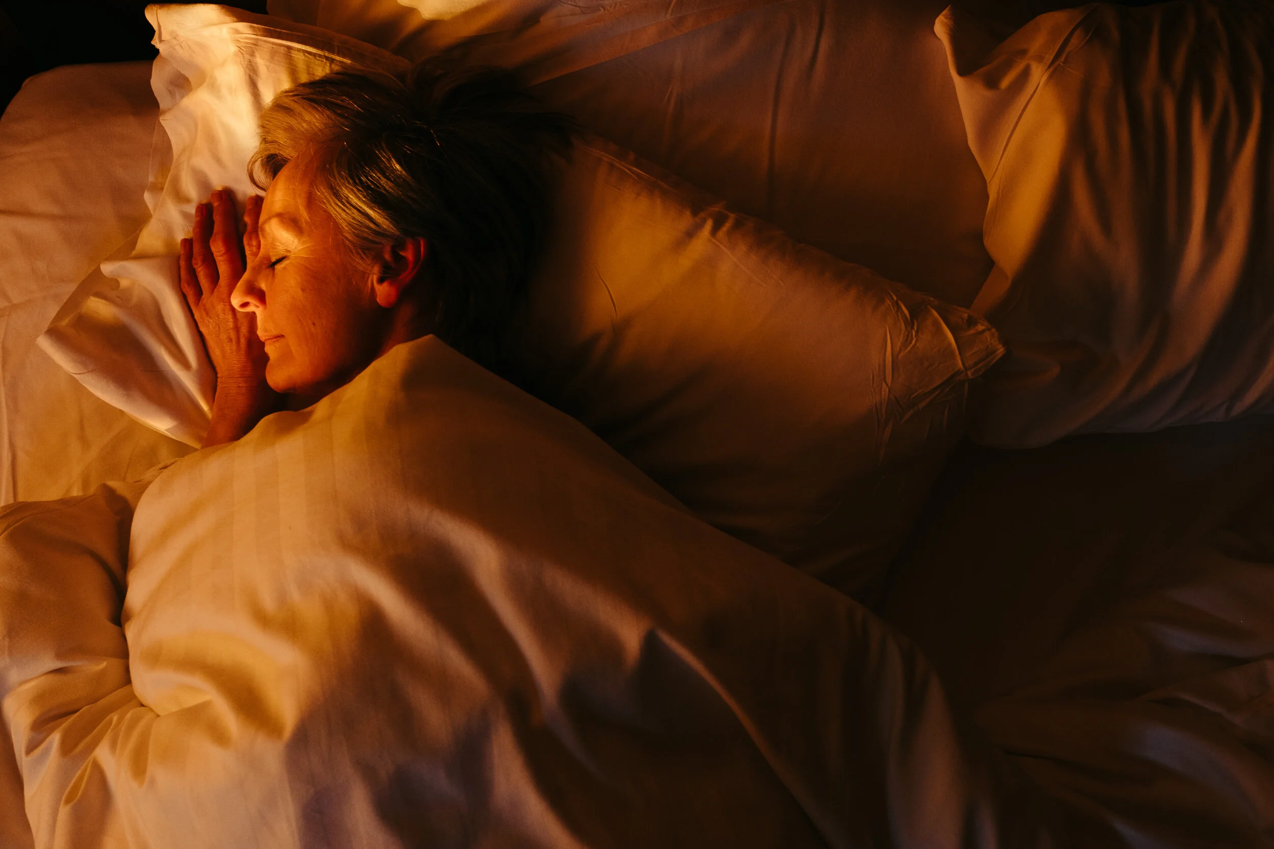 A woman with blonde hair lying in bed, resting her head on her hand with eyes closed, under warm lighting.