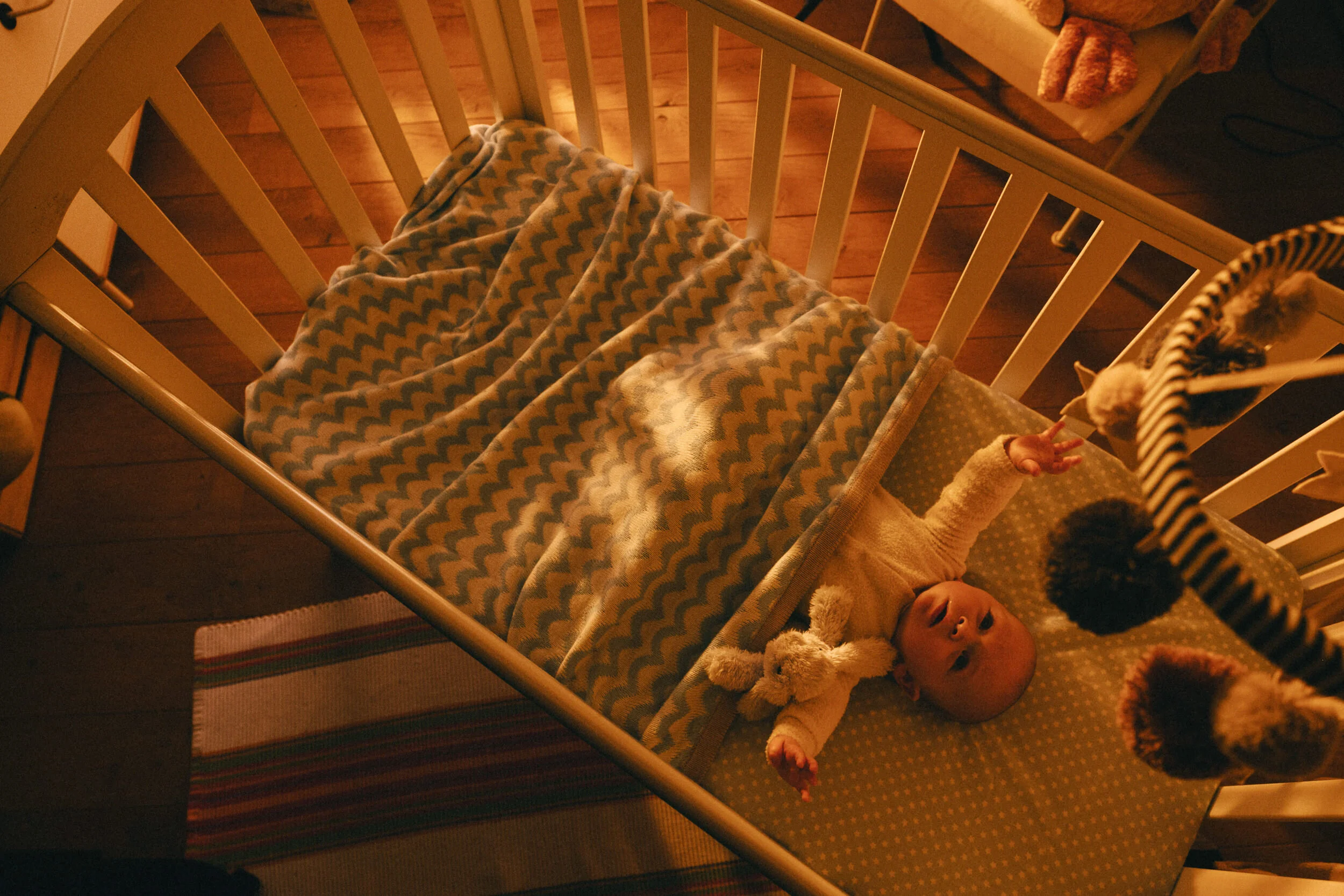 A baby lying in a crib, reaching up with one hand, wearing a yellow onesie with small polka dots, with a blanket covering part of their body. The crib has white slats and a stuffed toy or blanket at the head. The room has wooden flooring and warm lig