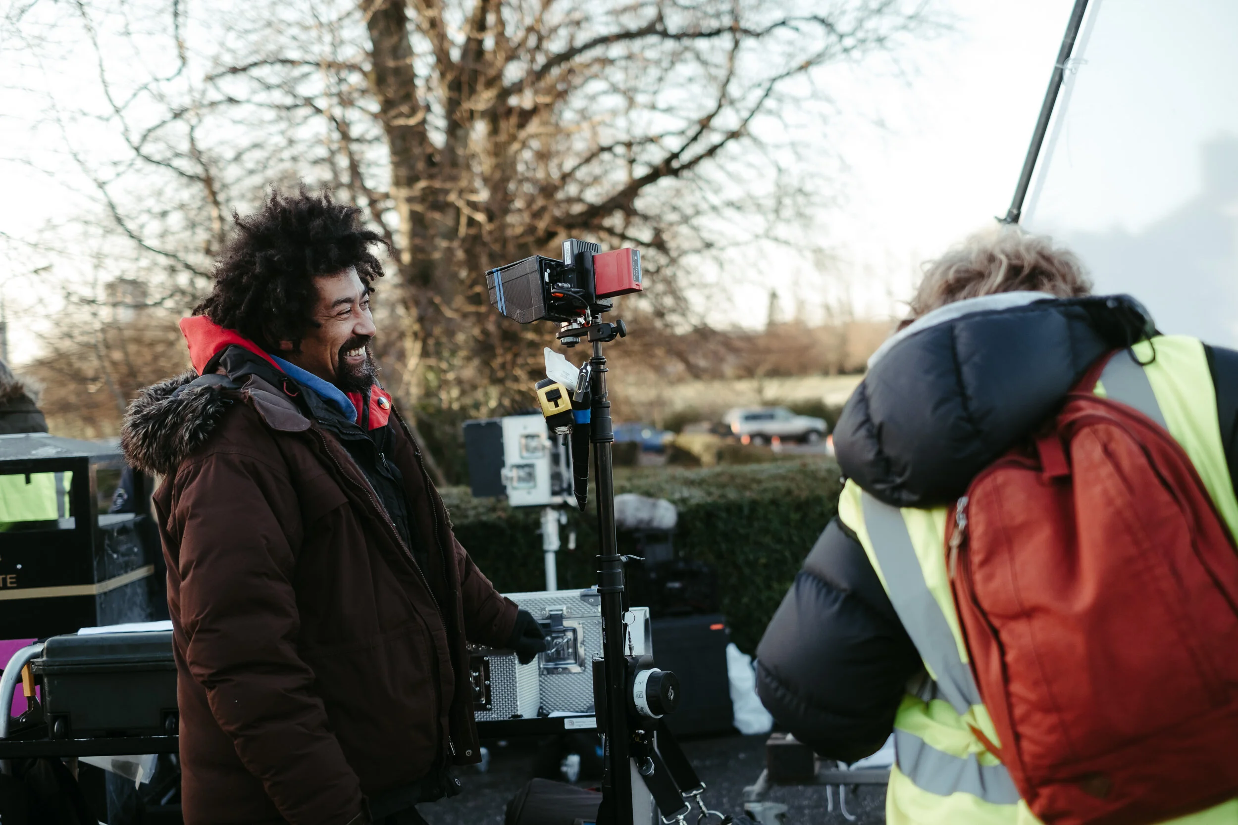 Two people on a film set outdoors, one smiling and wearing a brown jacket with a fur-lined hood, the other with gray hair in a neon yellow safety vest and red backpack, standing near camera equipment and a large video monitor, with leafless trees in 