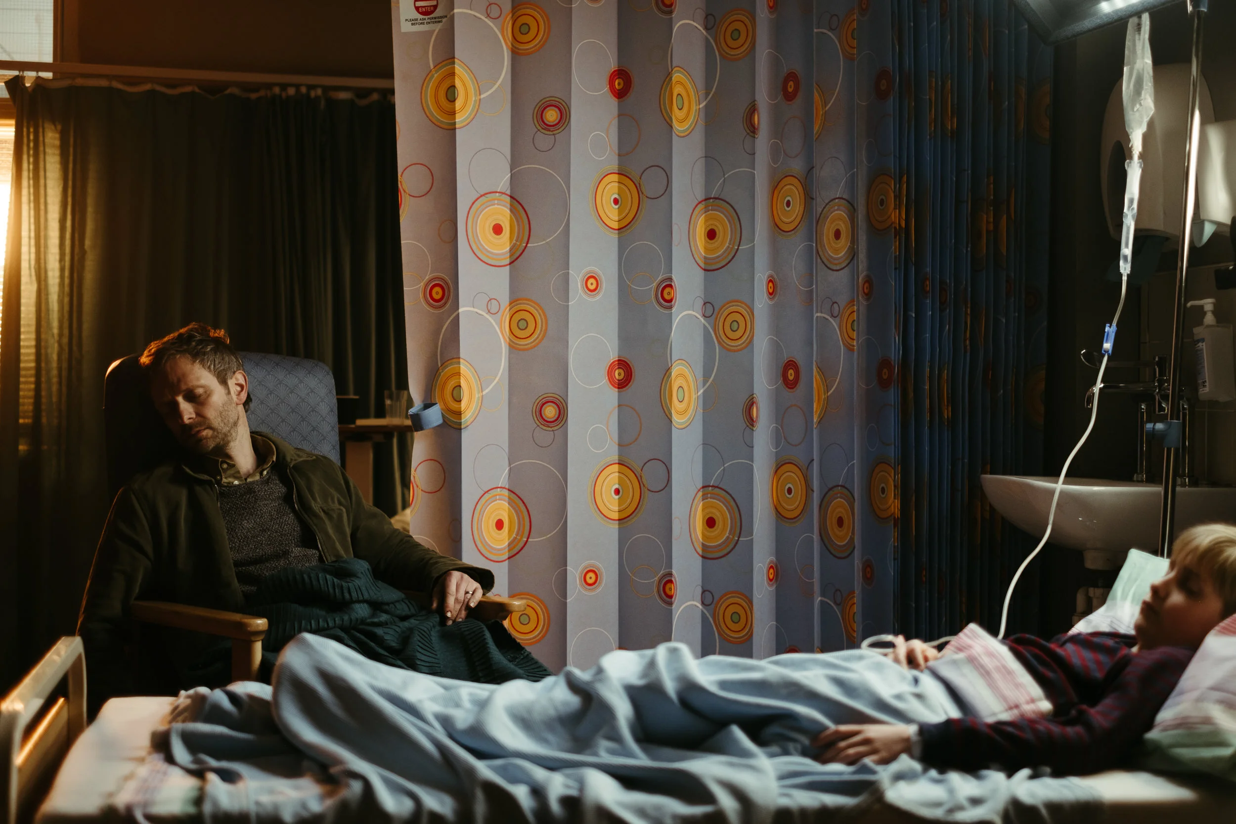 A man sleeping on a hospital bed while a child lies on a bed nearby, with colorful circular patterned curtains in the background and medical equipment on the side.