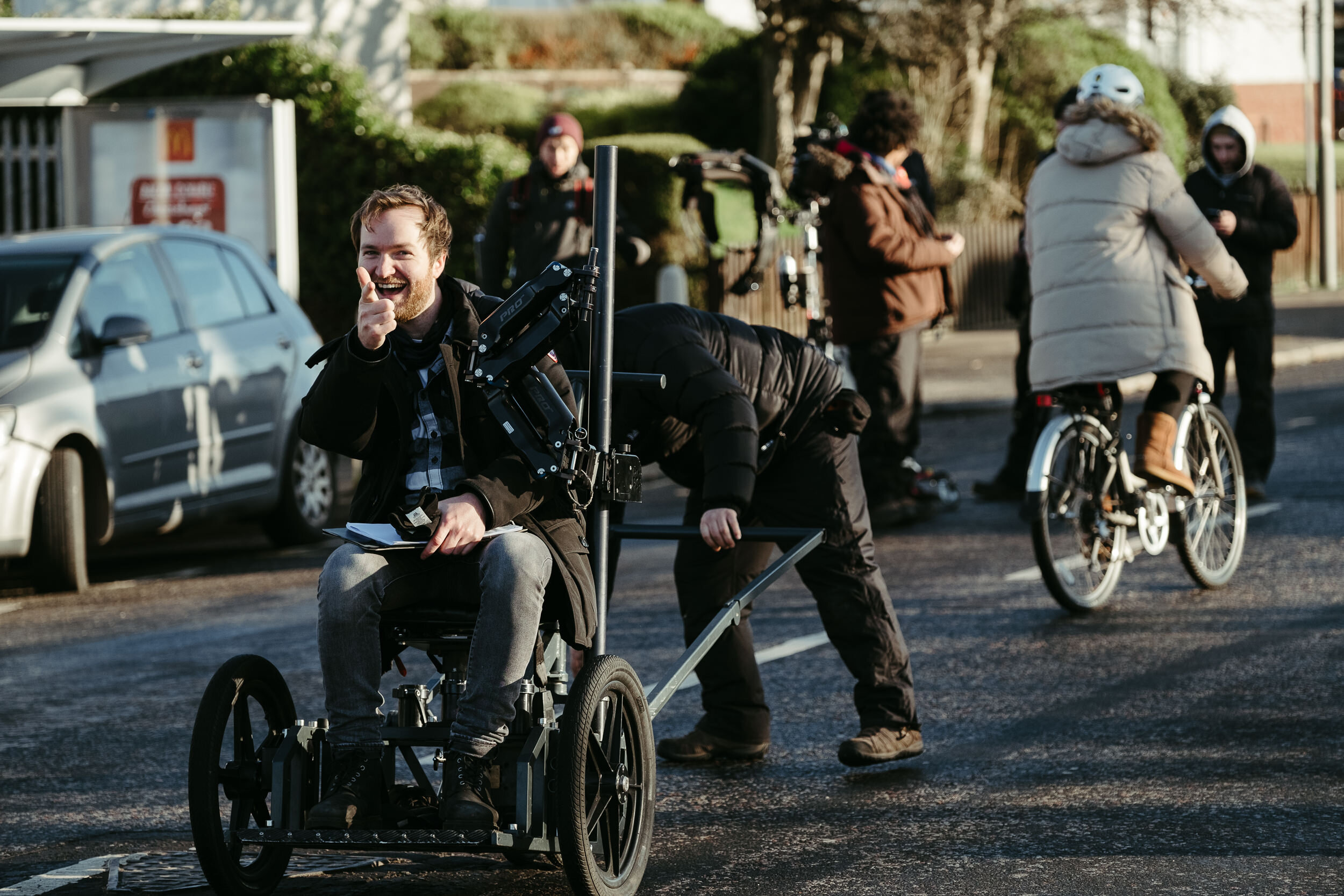 A man in a wheelchair smiling and pointing, with others standing, biking, and filming nearby in an outdoor urban setting.
