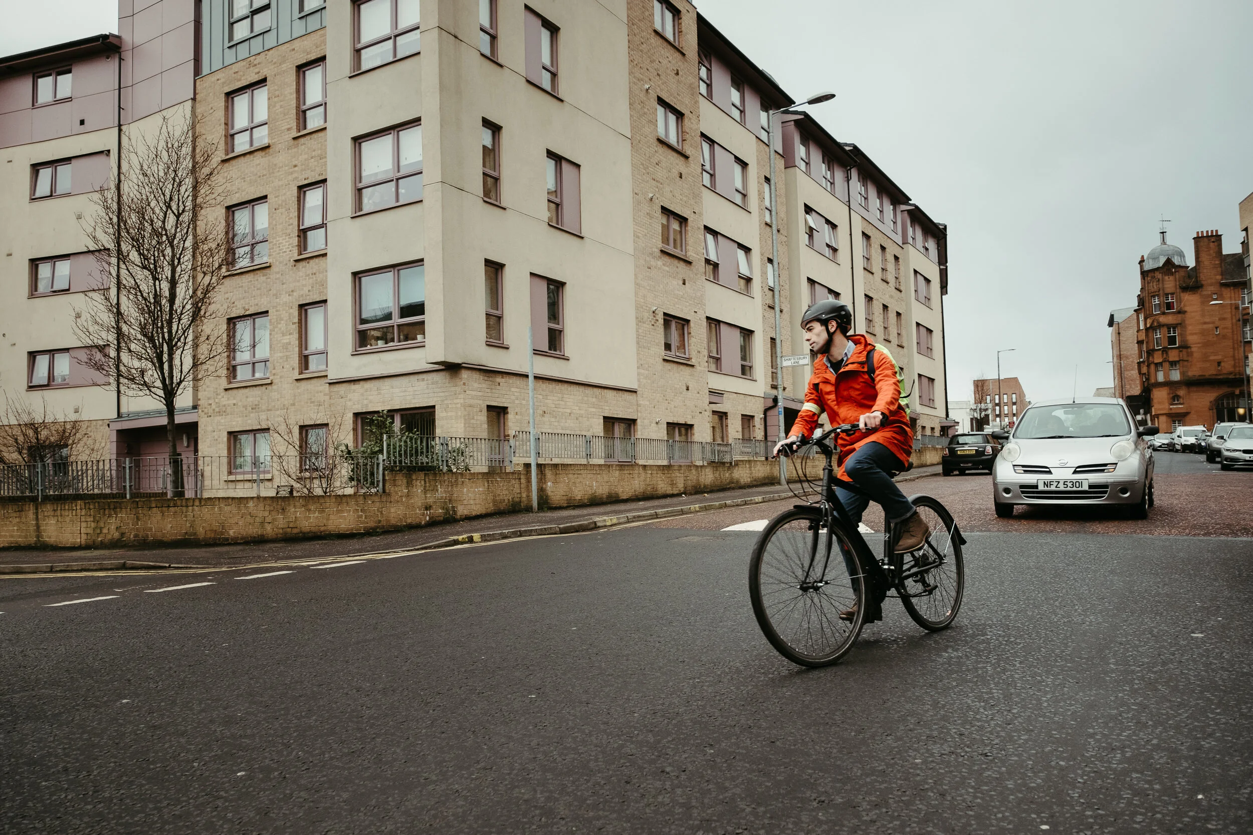 A man wearing an orange jacket and black helmet riding a bicycle on a city street, with cars parked on the side and a multi-story residential building in the background.