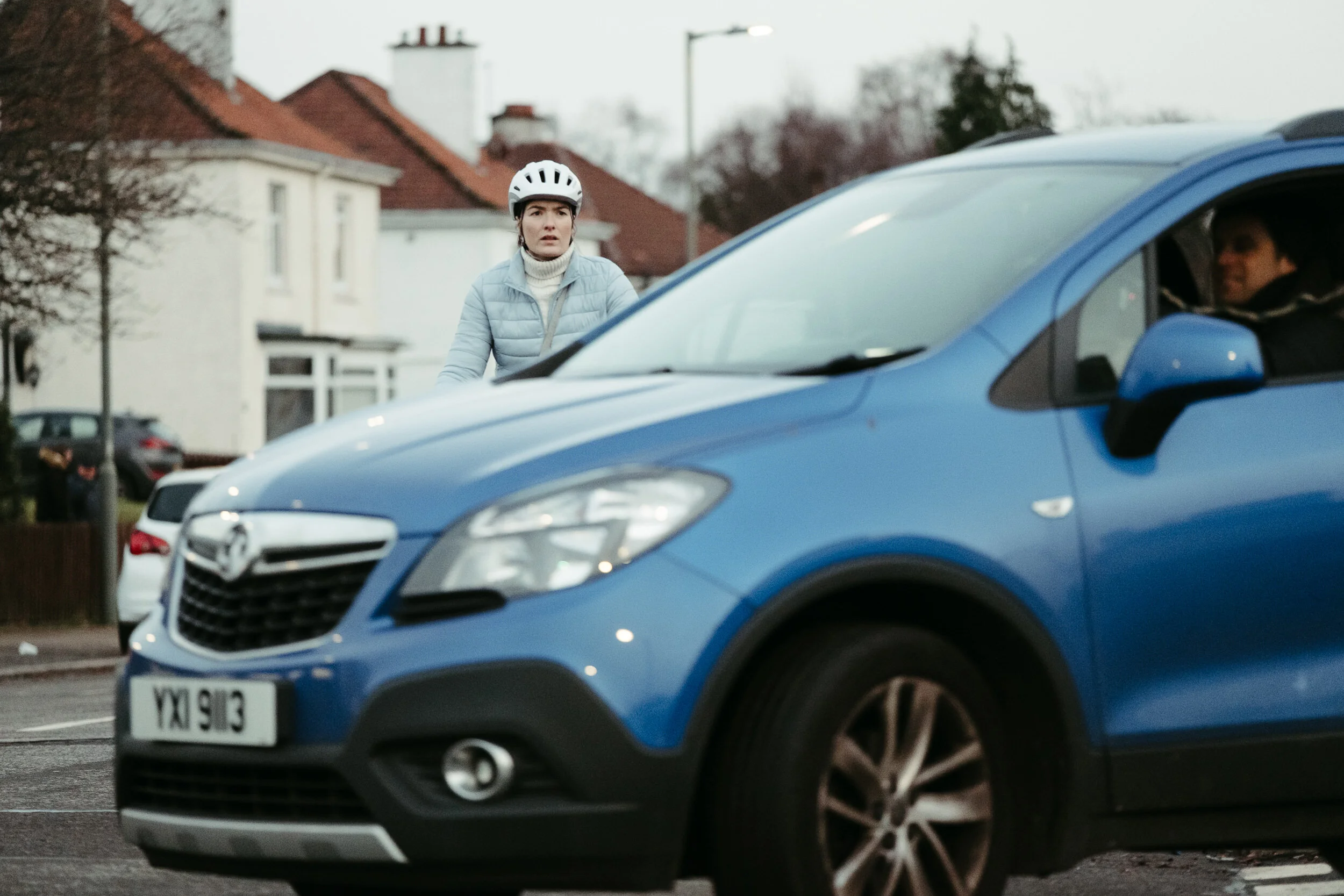 A woman wearing a white helmet and a light blue puffer jacket, looking surprised or concerned, standing outdoors near a row of residential houses, with a blue car in the foreground and a man visible inside the car.