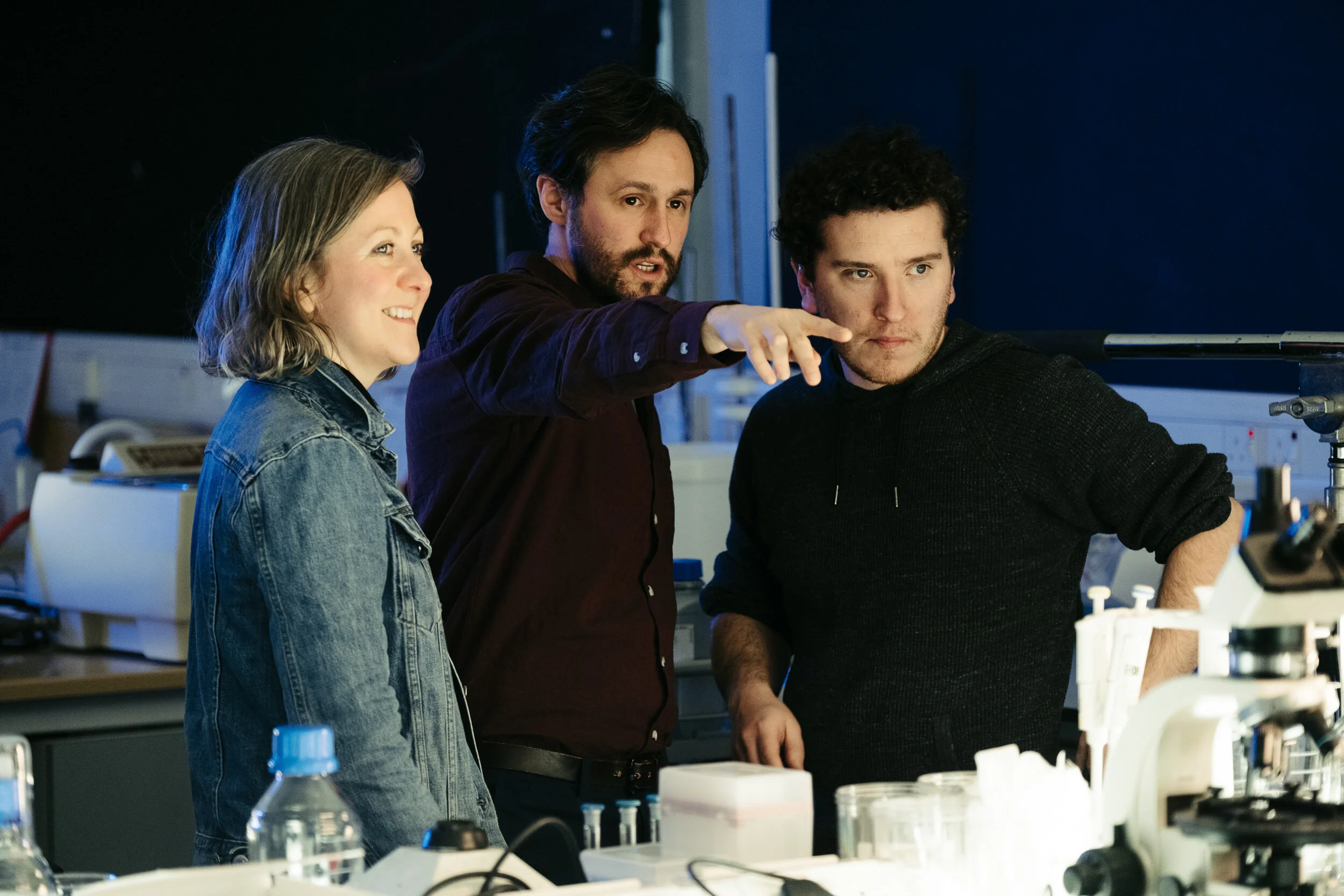 Three scientists, two men and one woman, in a laboratory analyzing scientific data or experiments, with laboratory equipment and supplies around them.