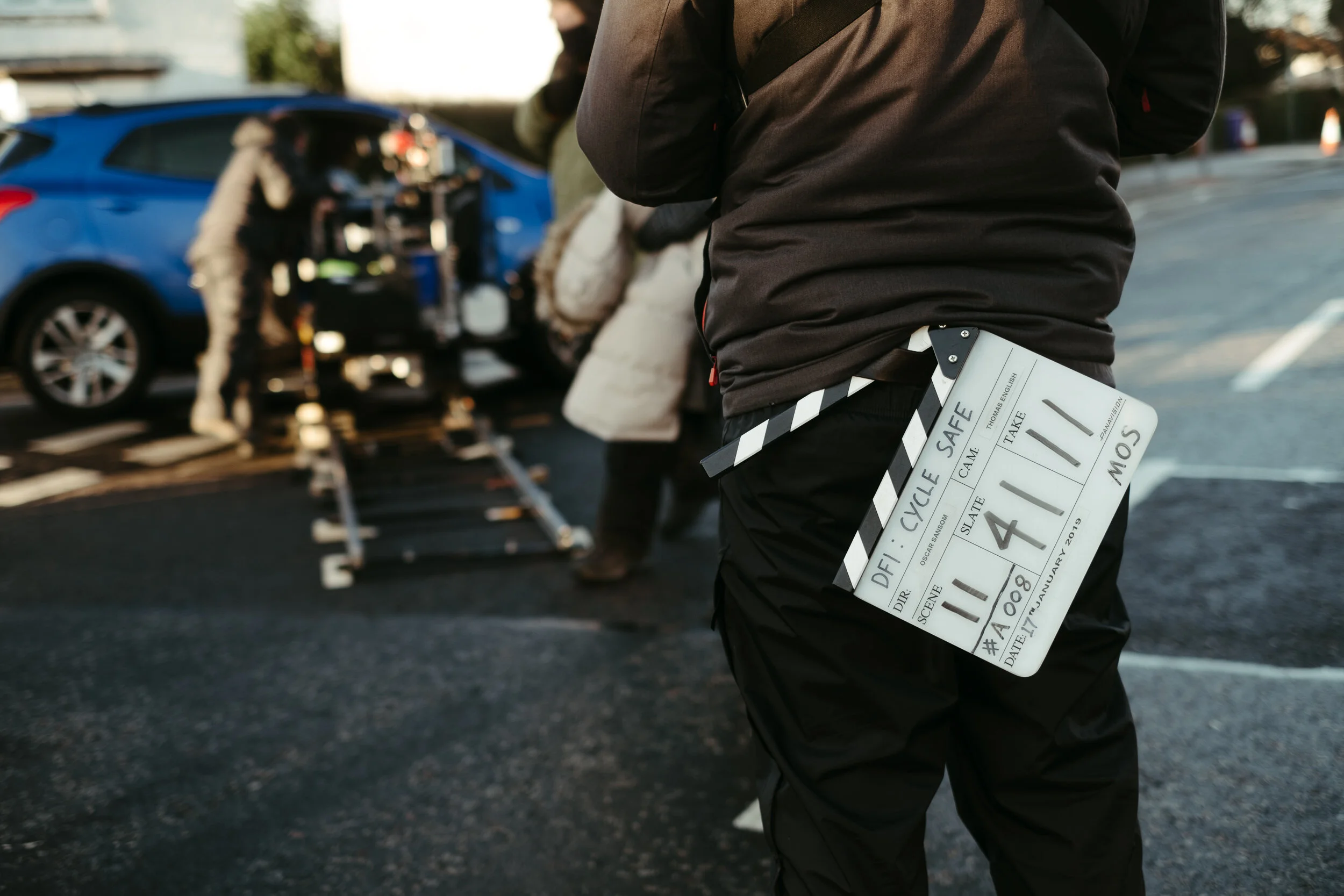 A person dressed in black with a clapperboard attached to their waist holding a film slate with information about a scene being filmed on a parking lot. In the background, there are crew members and a blue car with its trunk open, possibly for filmin