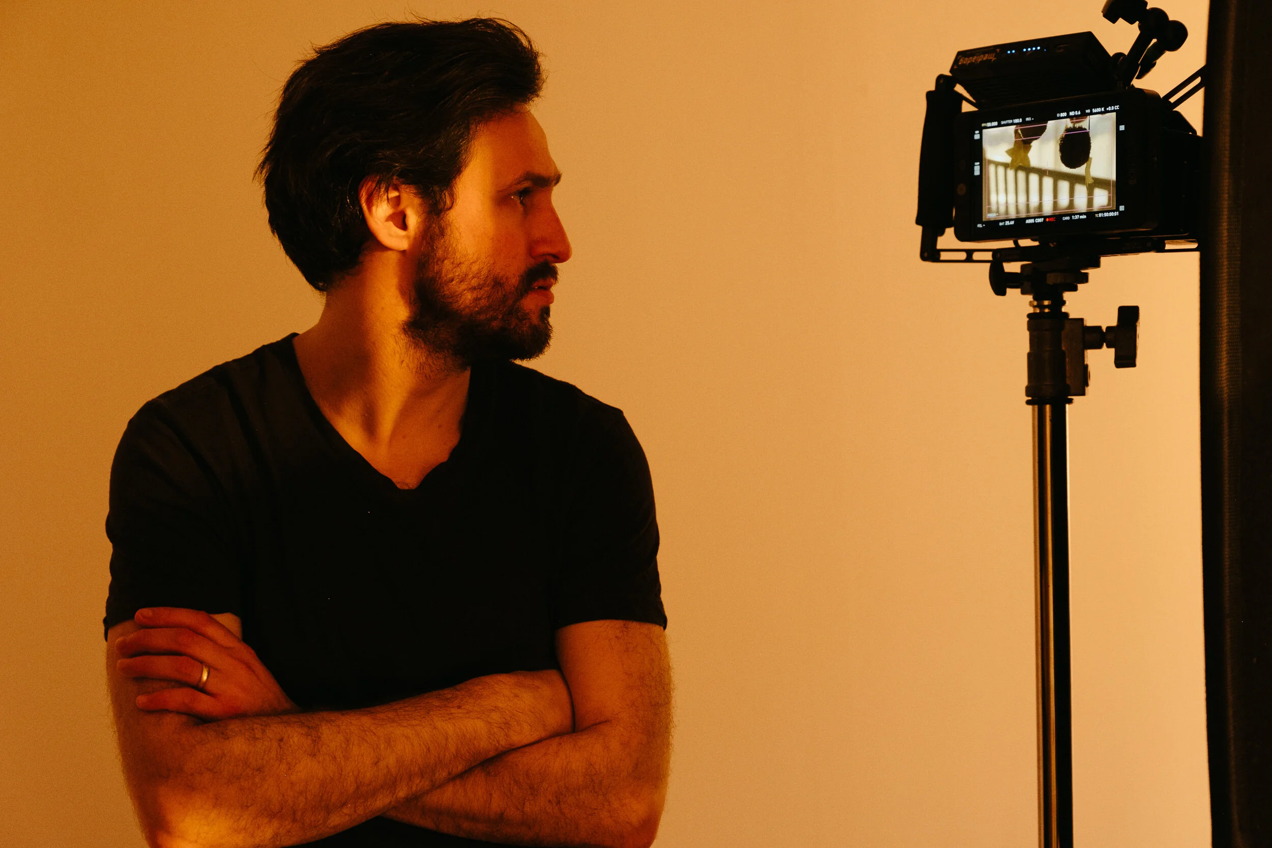 A man with dark hair and a beard stands in profile with crossed arms, facing a professional video camera on a tripod, against a plain beige background.