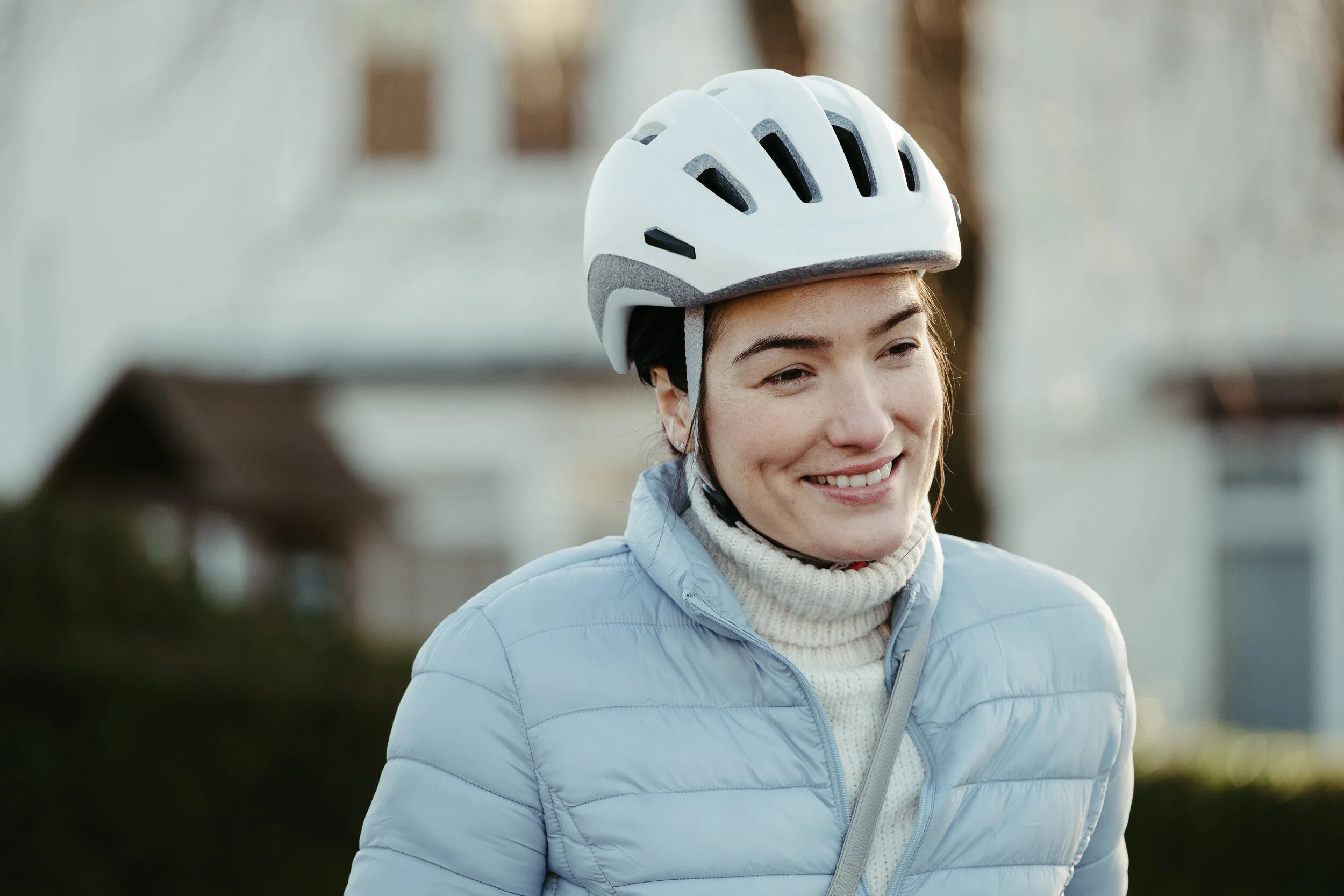 A woman smiling outdoors wearing a white bike helmet and light blue puffer jacket.