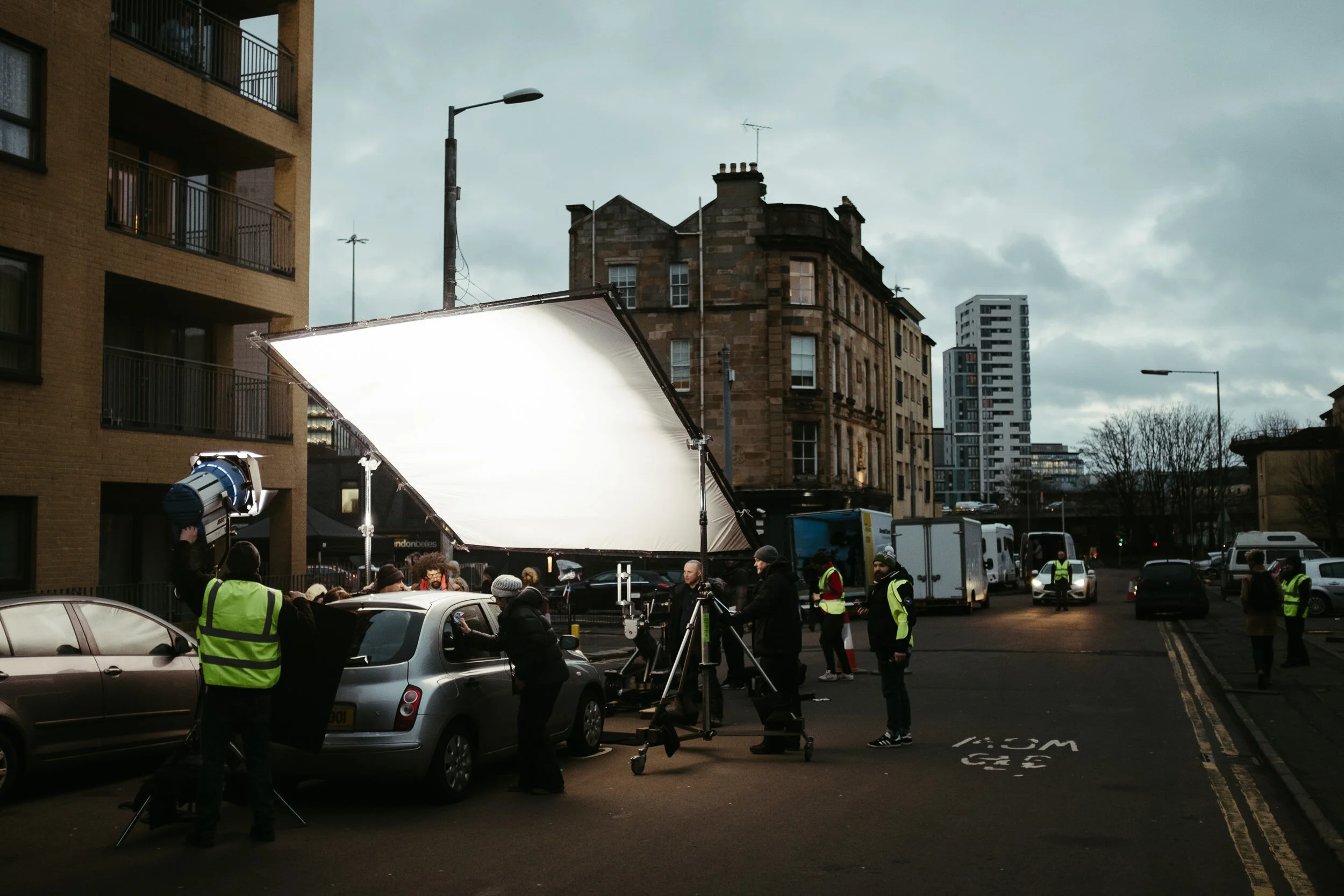 Filming crew setting up equipment on a city street with buildings, cars, and cloudy sky in the background.