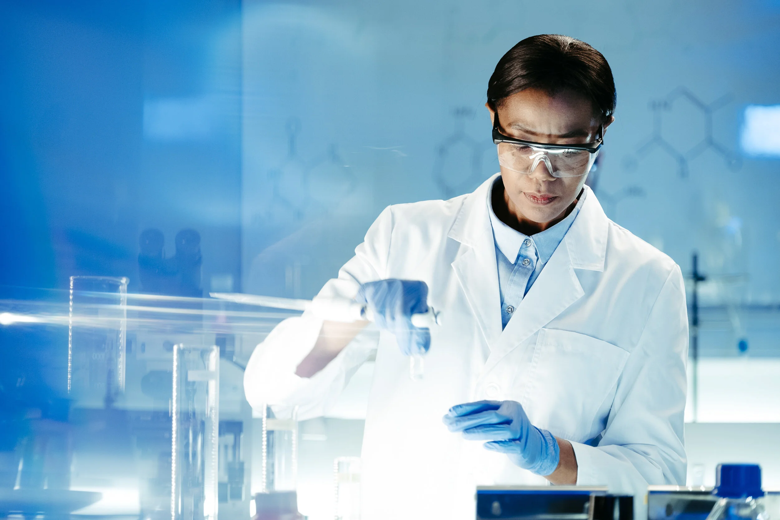 A scientist wearing safety goggles, gloves, a white lab coat, and a blue shirt works with laboratory equipment in a modern, glass-walled laboratory.