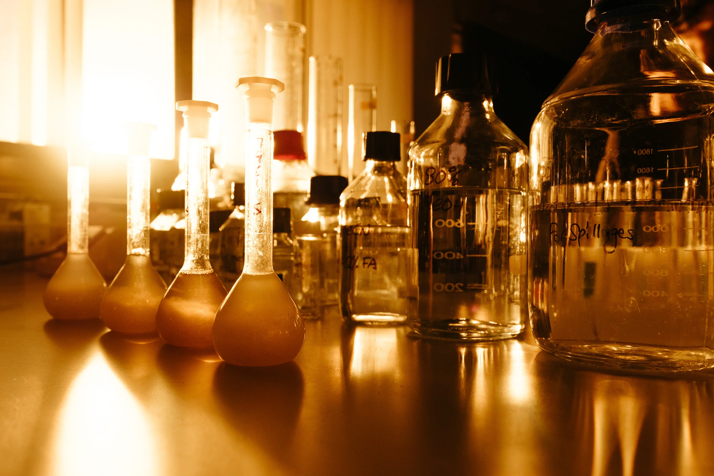 A close-up of various laboratory glass bottles and test tubes filled with liquids, positioned on a reflective surface with sunlight streaming in from a window in the background.
