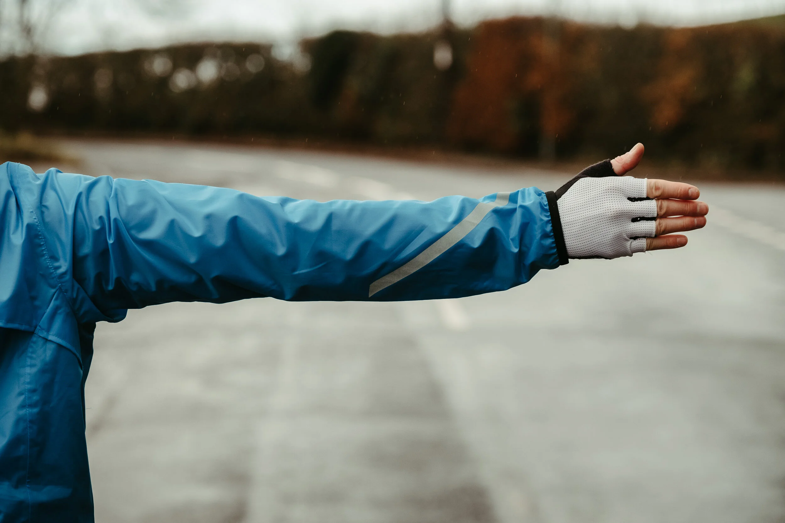 Close-up of a person wearing a blue jacket and gray gloves, extending their arm outward in an outdoor setting with an overcast sky and autumn trees in the background.