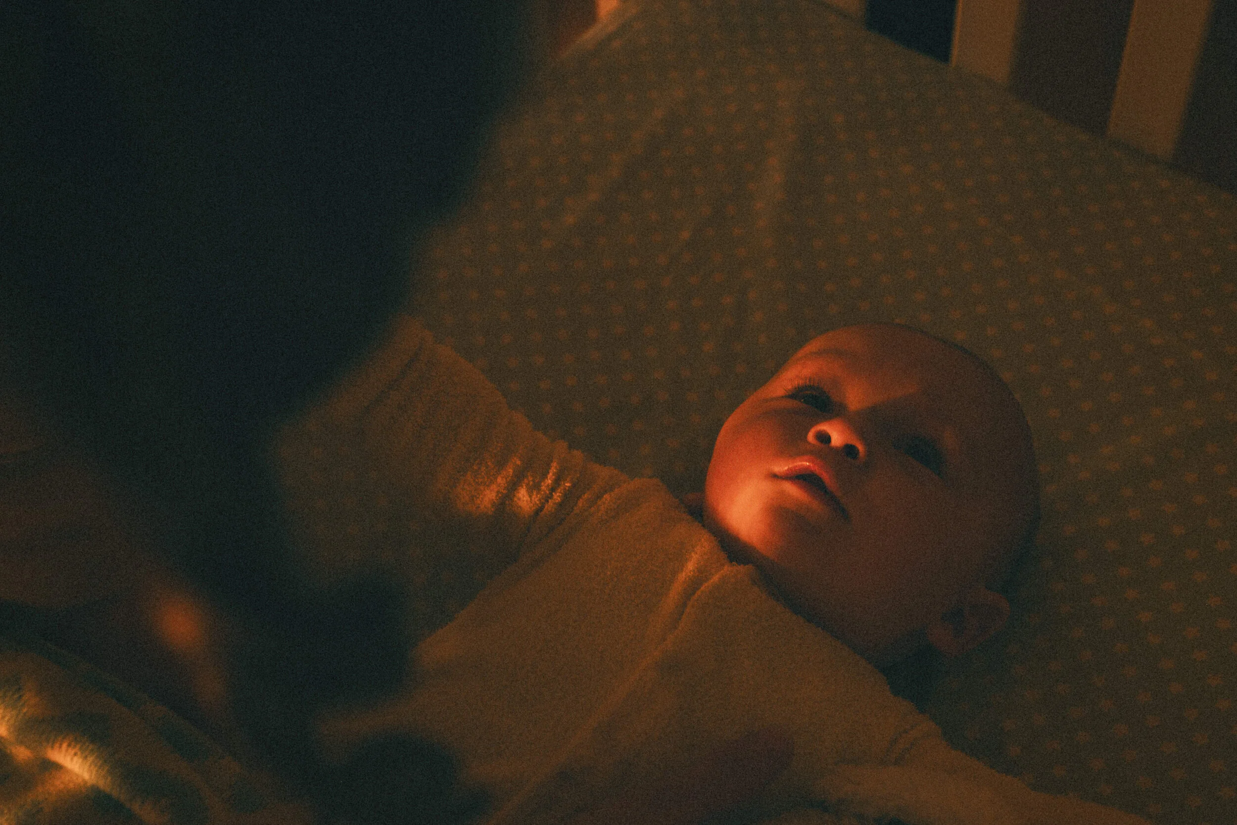 A baby lying on the floor with a blanket over their body, looking up at the camera, with a warm light illuminating their face.