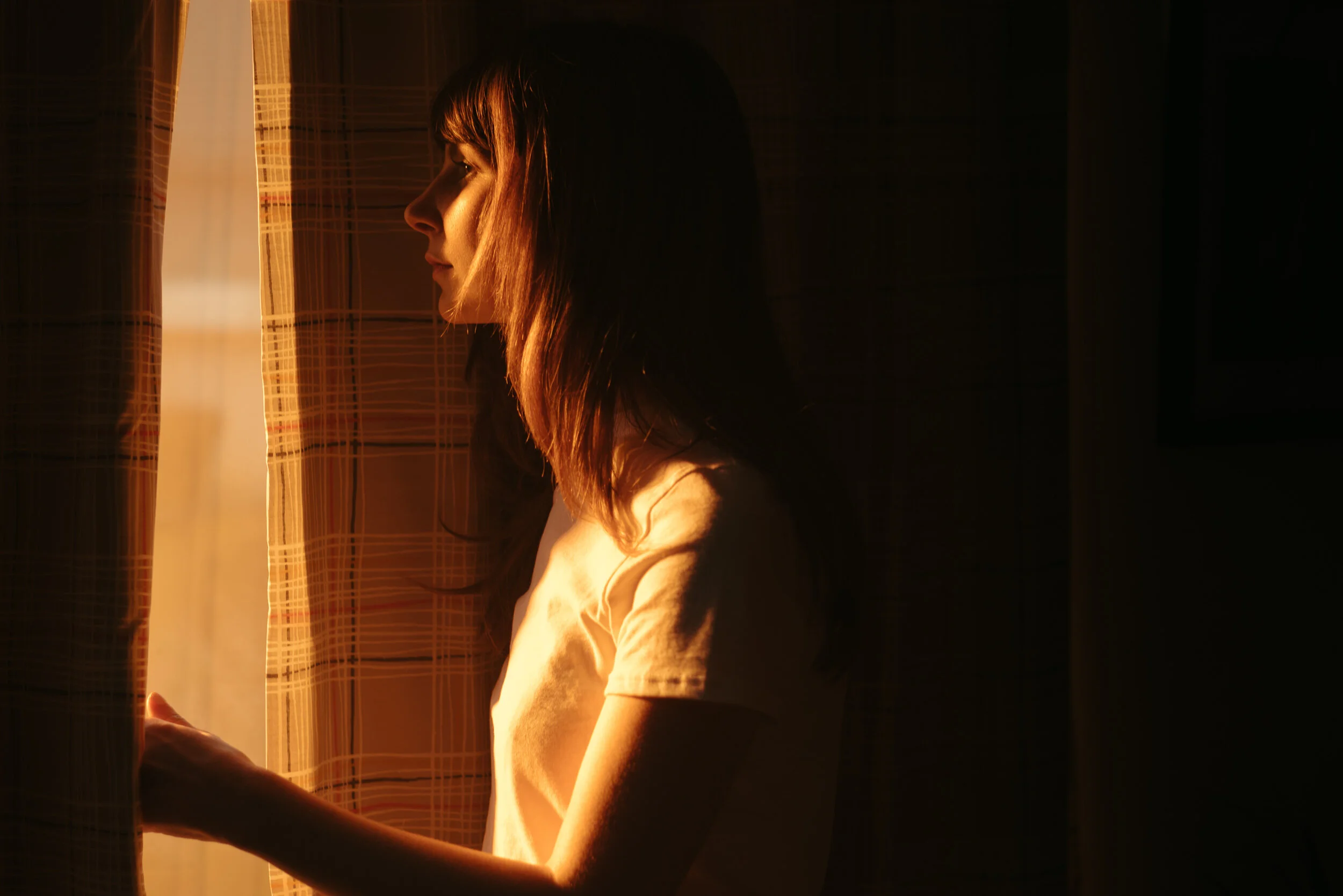 A young woman with light brown hair looking out a window at sunset, with sunlight illuminating her profile.