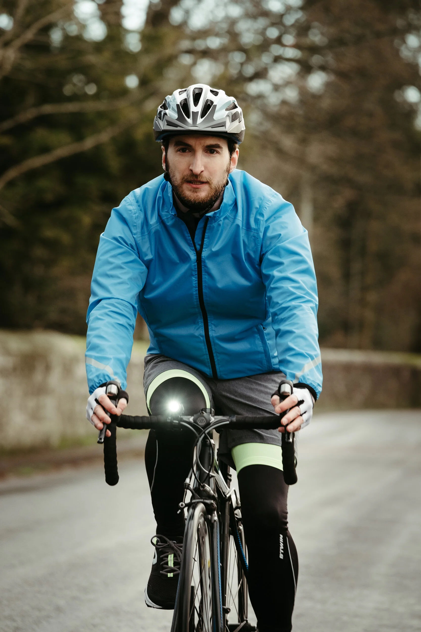 A male cyclist wearing a helmet and a blue jacket riding a bicycle on a paved path with trees in the background.