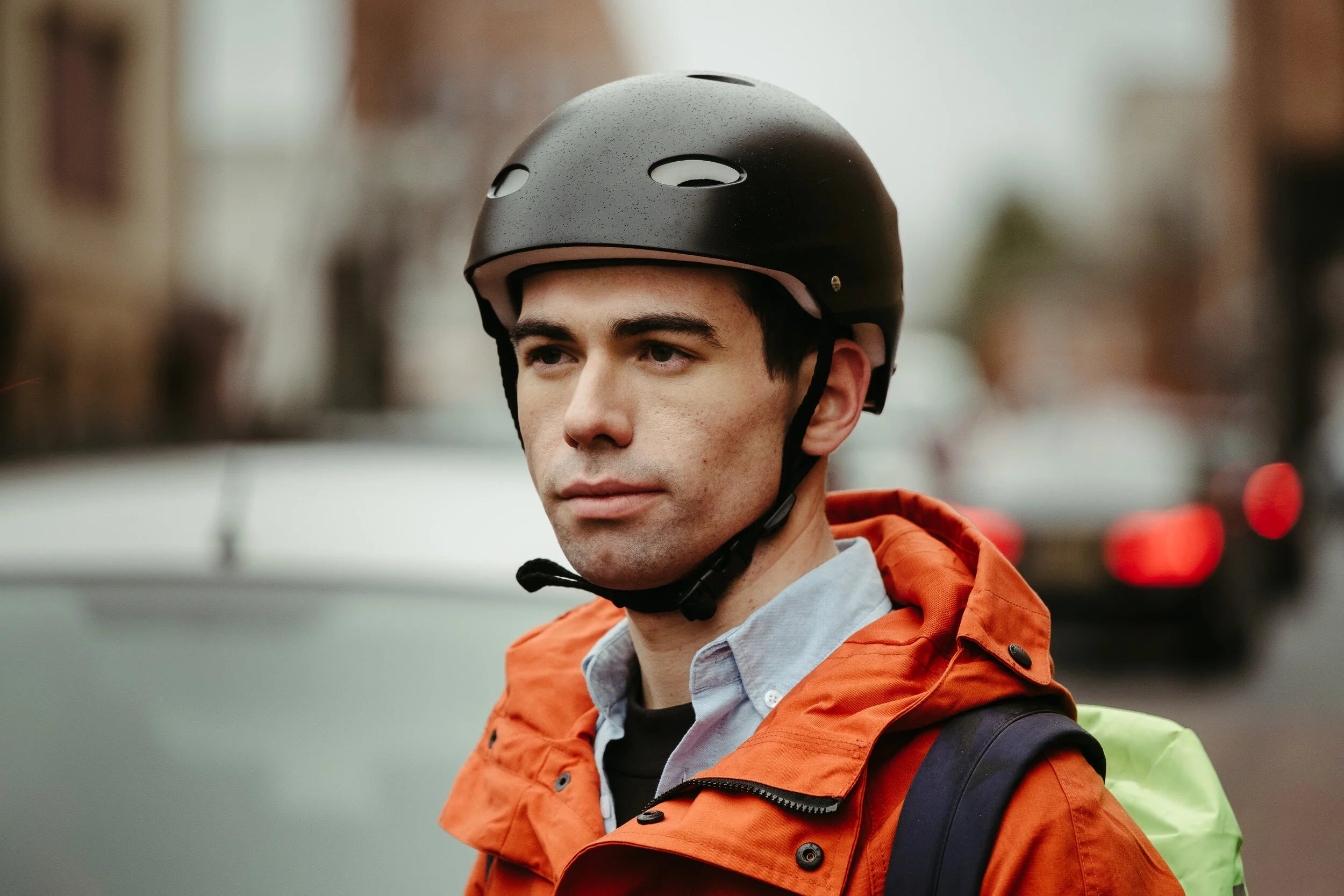 A young man wearing a black helmet, an orange jacket, and a backpack, standing outdoors with an out-of-focus background.