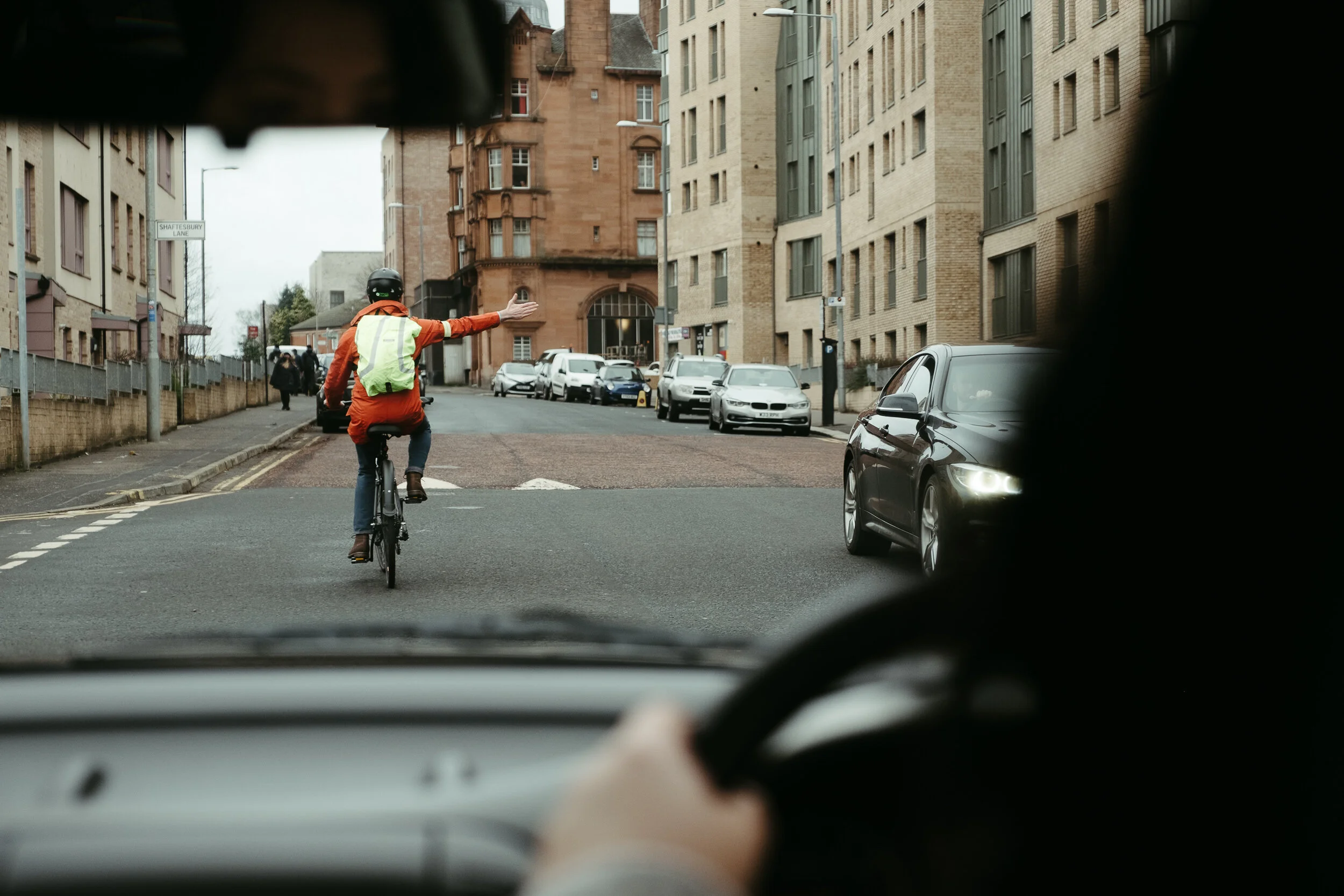 View from inside a car of a cyclist riding on the street, wearing an orange jacket, a green safety vest, and a black helmet, with buildings and parked cars on either side of the street.