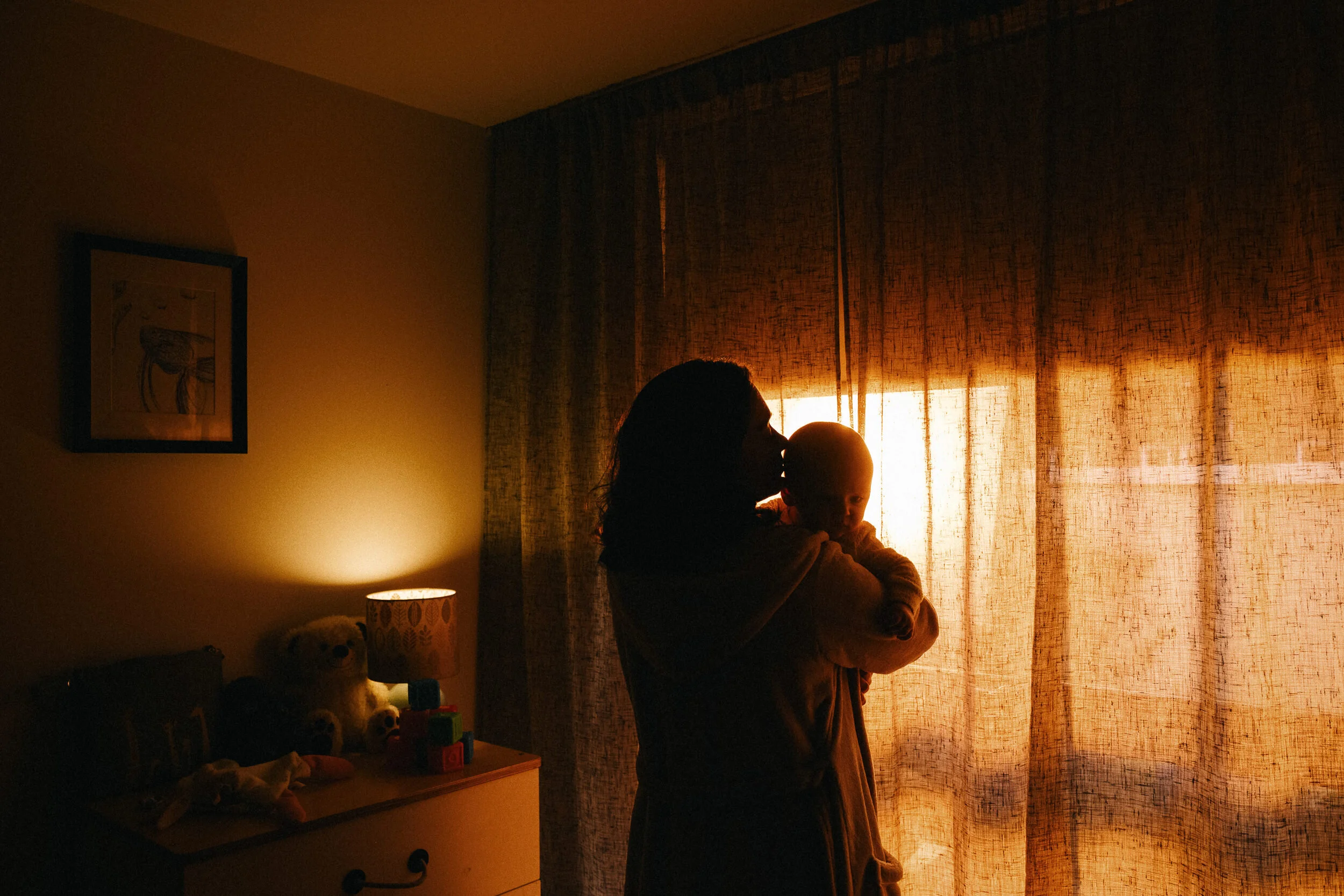 Silhouette of a woman holding a baby in front of a window with curtains, warm orange lighting in a dimly lit room.