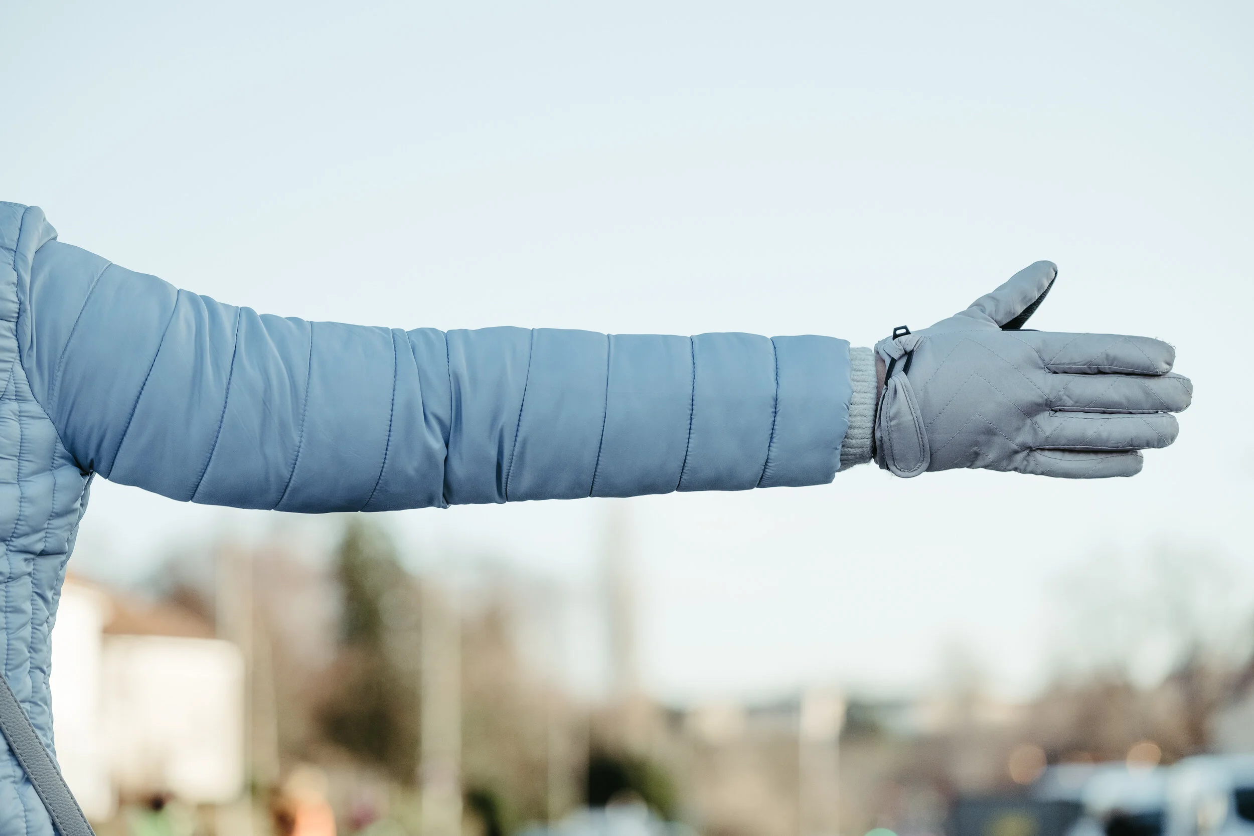 Person wearing a blue winter jacket and gray gloves giving a thumbs-up outdoors.