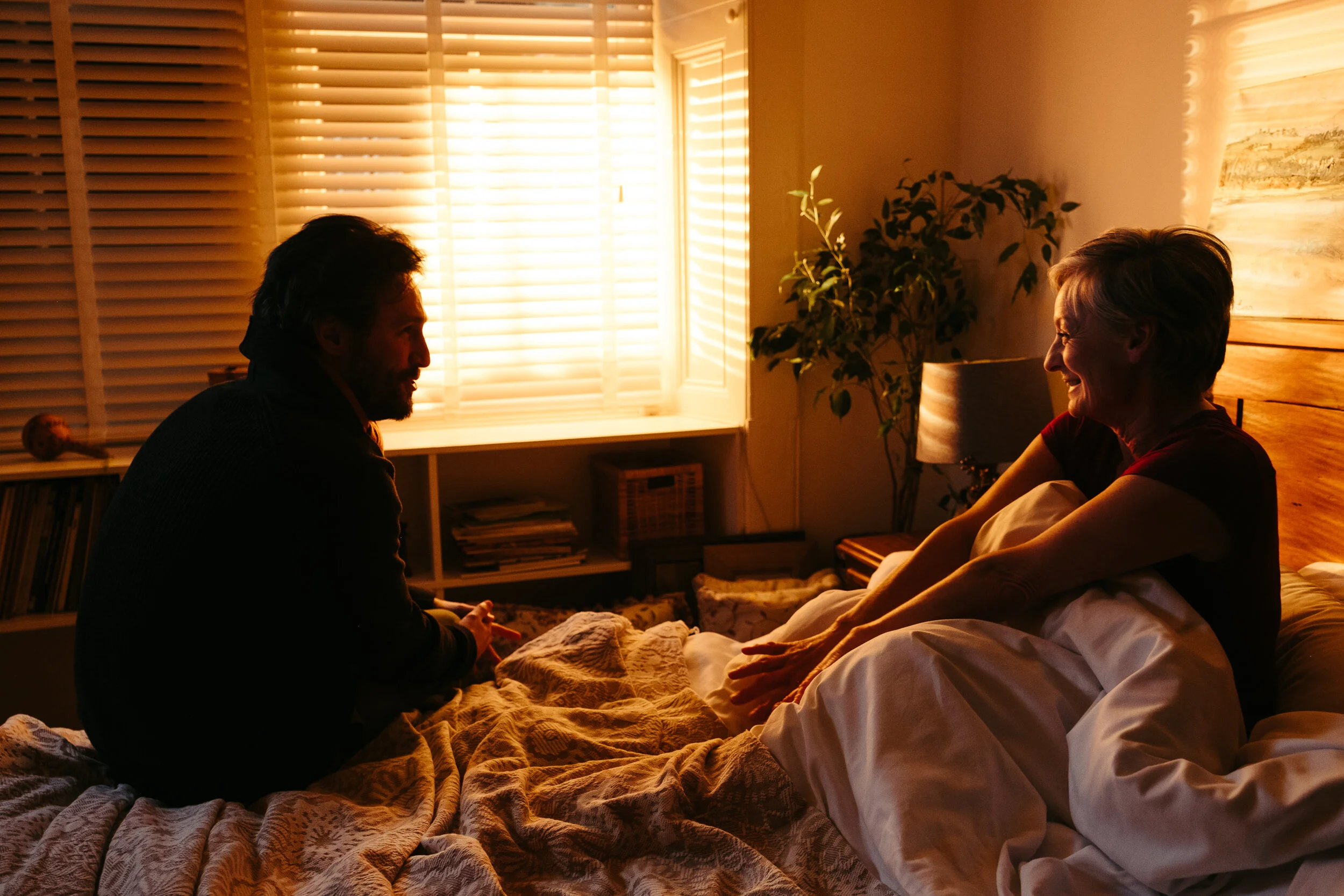 A man and an older woman sitting on a bed, facing each other, inside a warmly lit room with closed window blinds, a plant, and a painting on the wall.