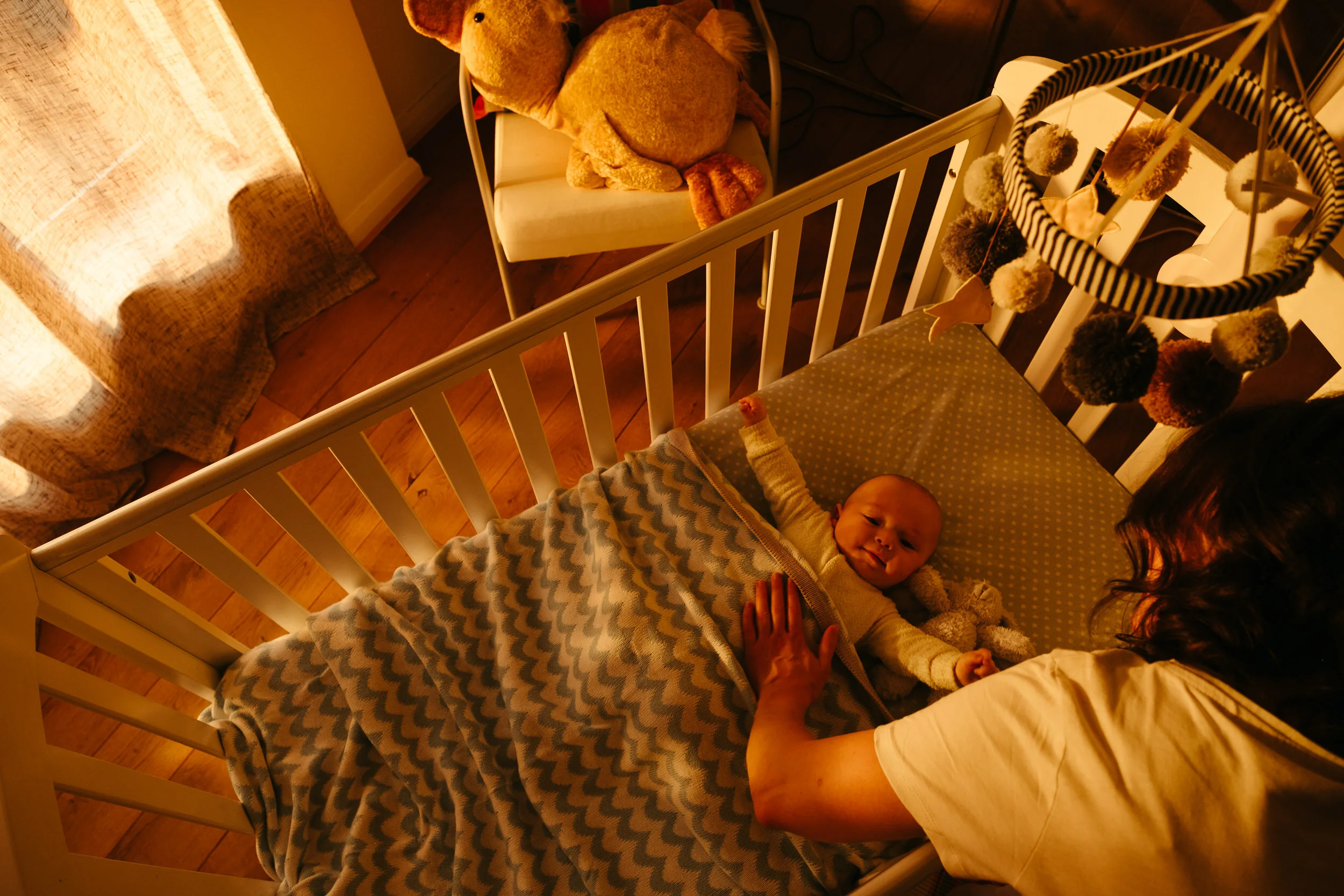 A baby lying in a crib with a woman holding the baby's hand. The crib has a blanket with a wave pattern. To the side, there is a stuffed animal and a hanging decoration with pom-poms.