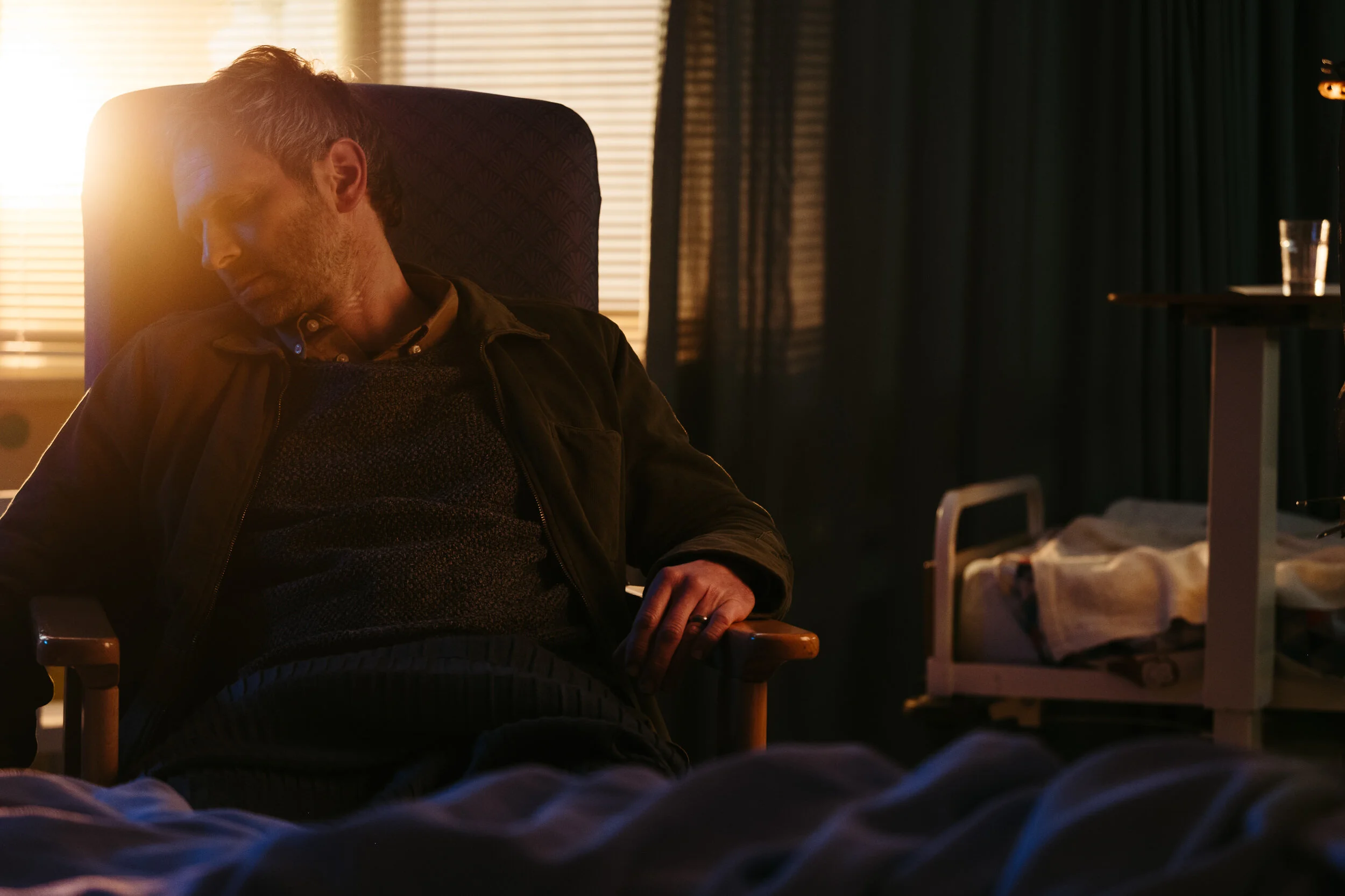 A man with light skin and short hair sleeping in a hospital bed with a head injury, wearing a dark jacket and patterned shirt, in a dimly lit room with sunlight coming through window blinds.