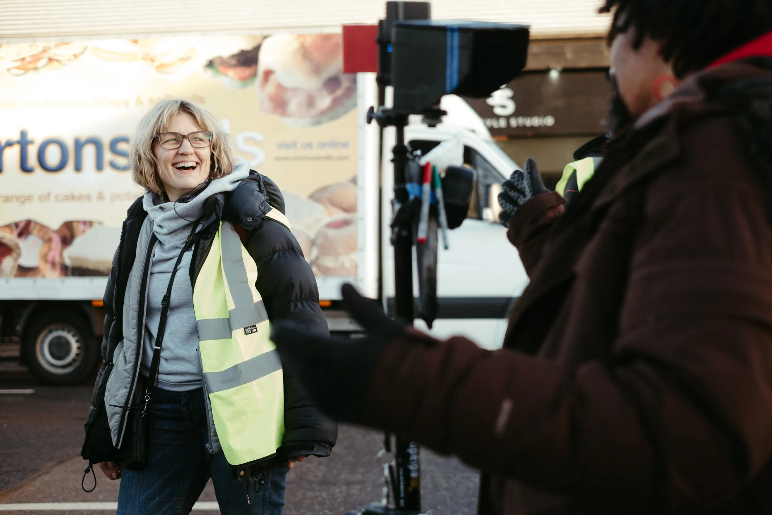 A woman with glasses smiling and talking to a person on a motorcycle, outdoors near a food truck. The woman is wearing a black jacket and a reflective safety vest.
