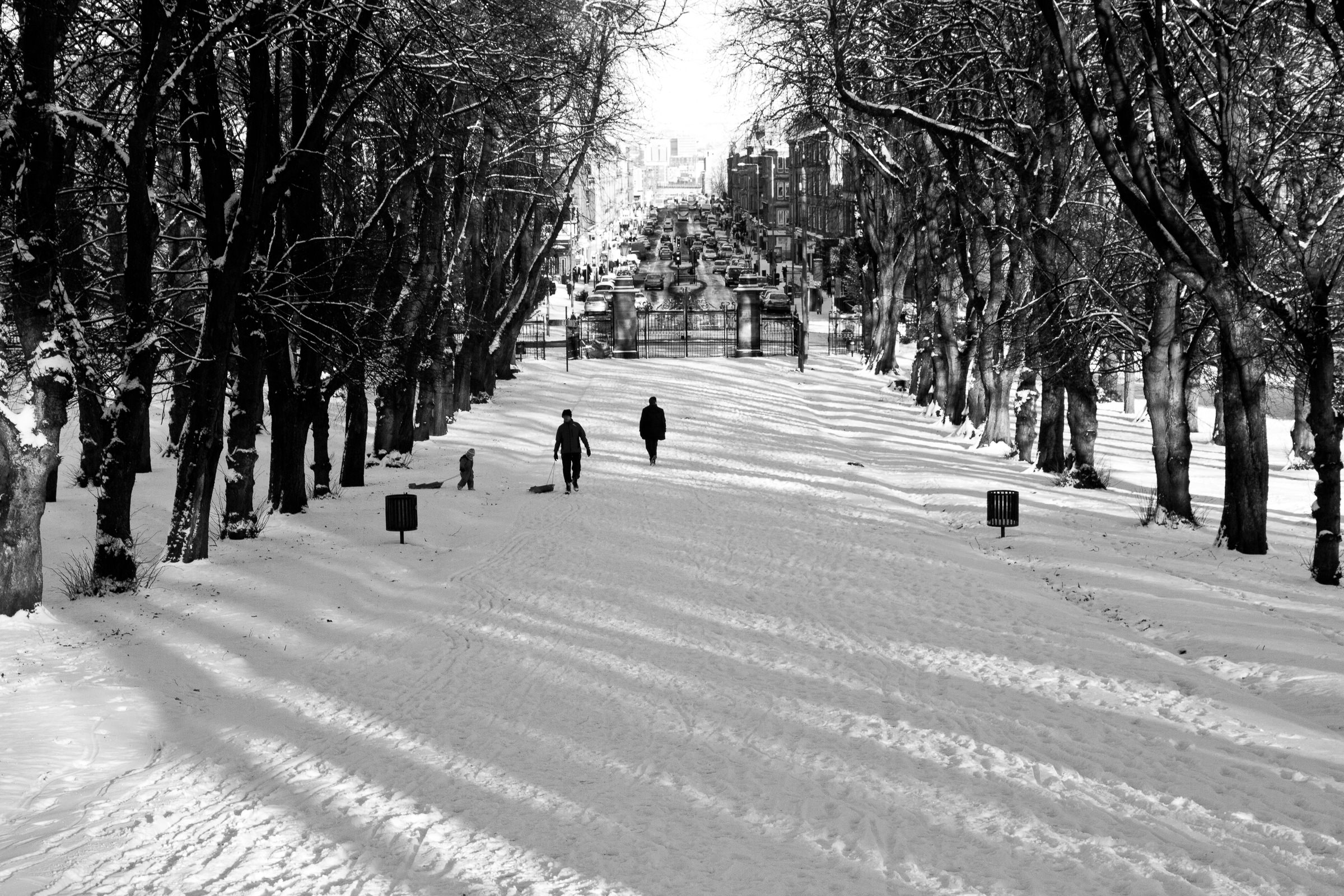 Snow-covered park with trees lining the path, three people walking, and a city street with cars in the background.