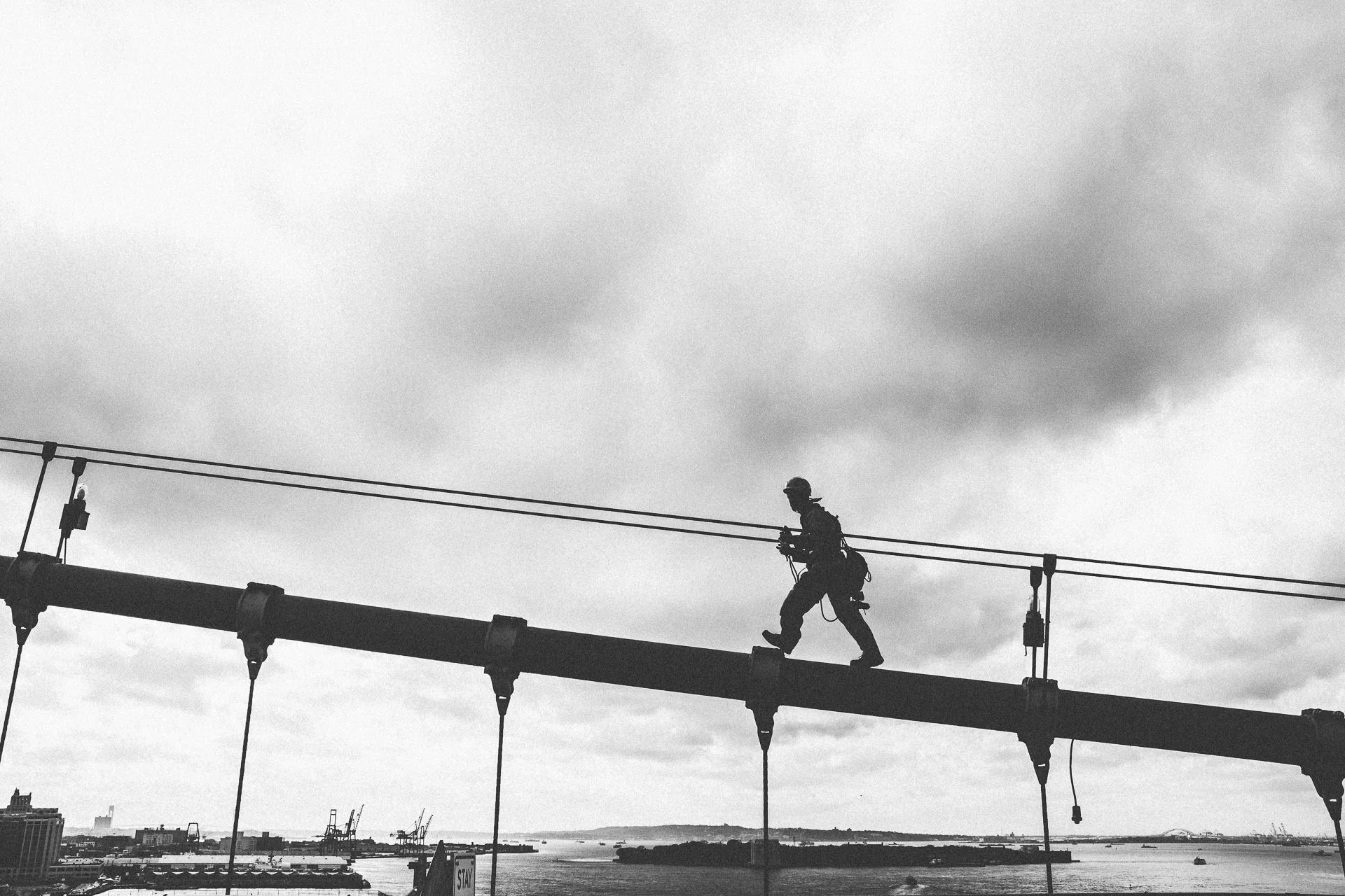 A person wearing a helmet and harness crossing a suspension bridge over a body of water with a cityscape and hills in the background, under cloudy skies.