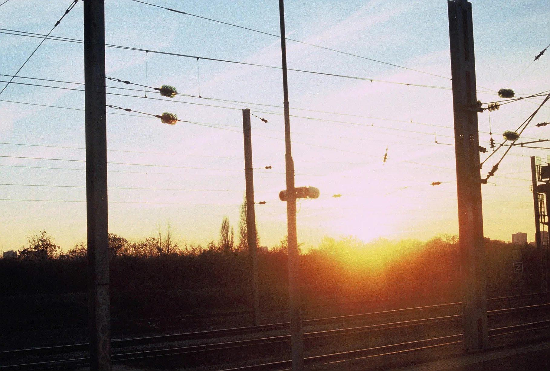 Sunset views of railroad power lines and poles along the tracks with trees and a building visible in the distance.