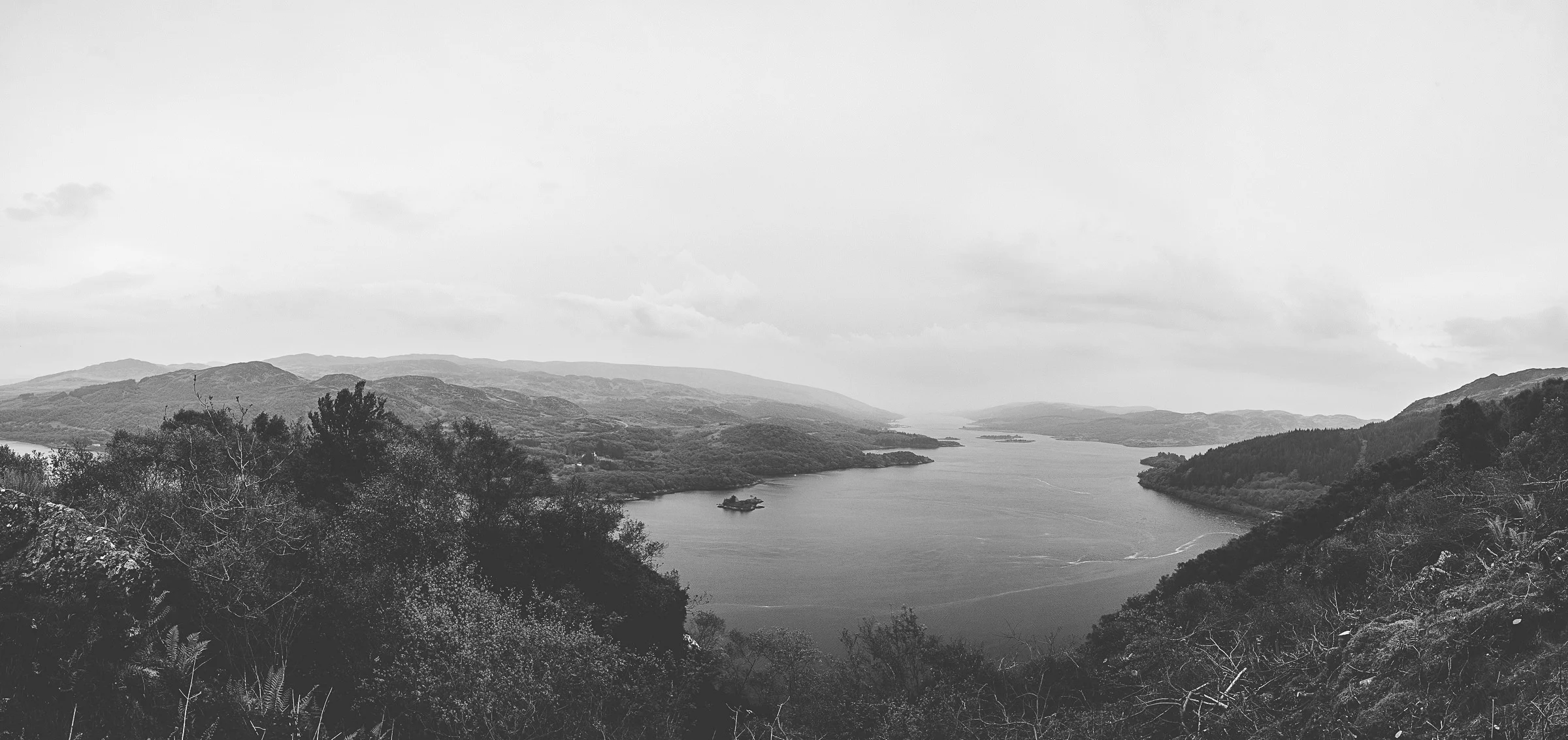 Black and white photograph of a river flowing between hills and mountains with trees in the foreground.
