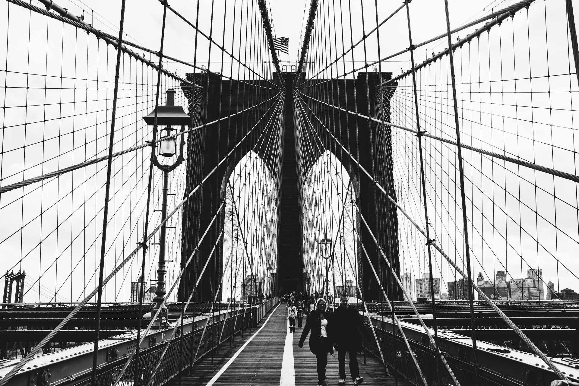 Black and white photograph of the Brooklyn Bridge in New York City, showing cables and the stone tower with an American flag on top, with people walking on the pedestrian walkway.