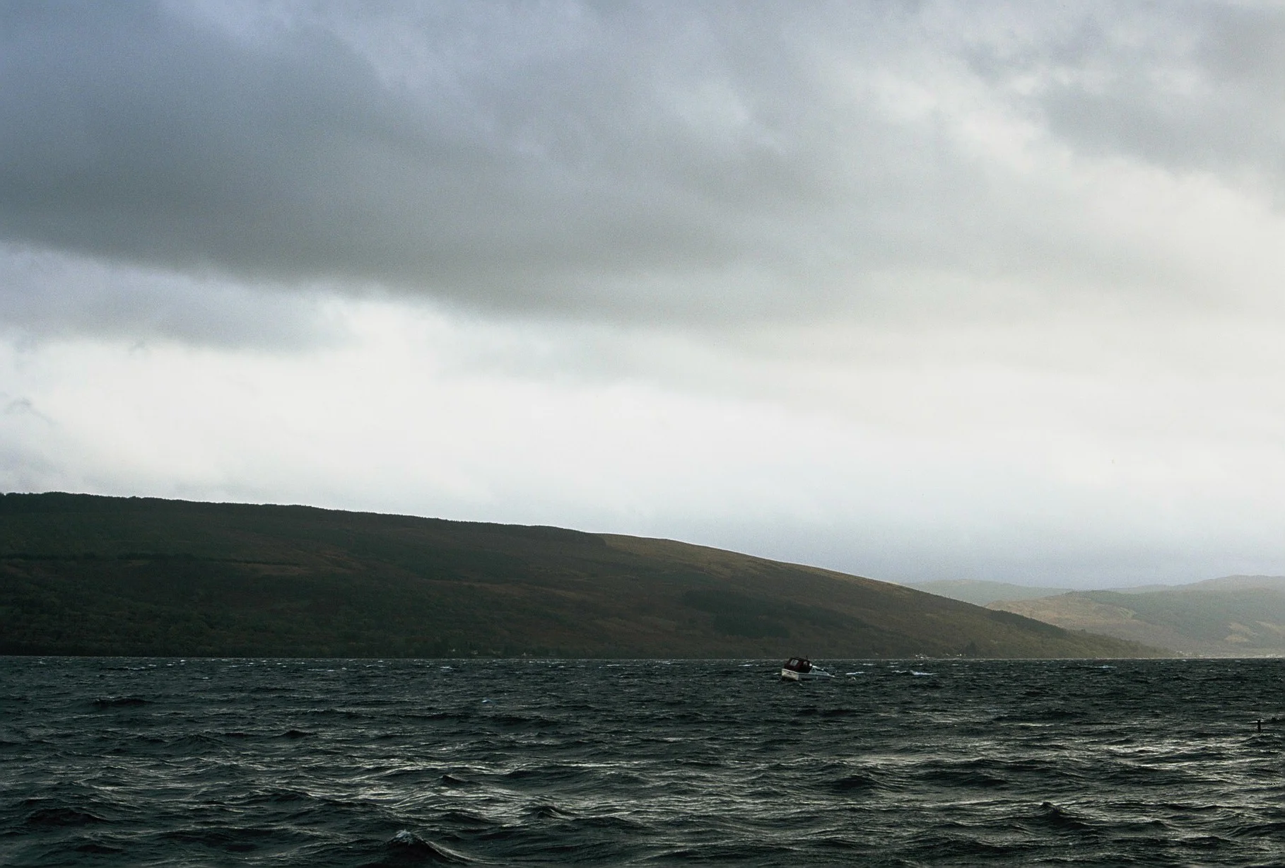 A boat on dark waters under a cloudy sky with a hilly, green landscape in the background.