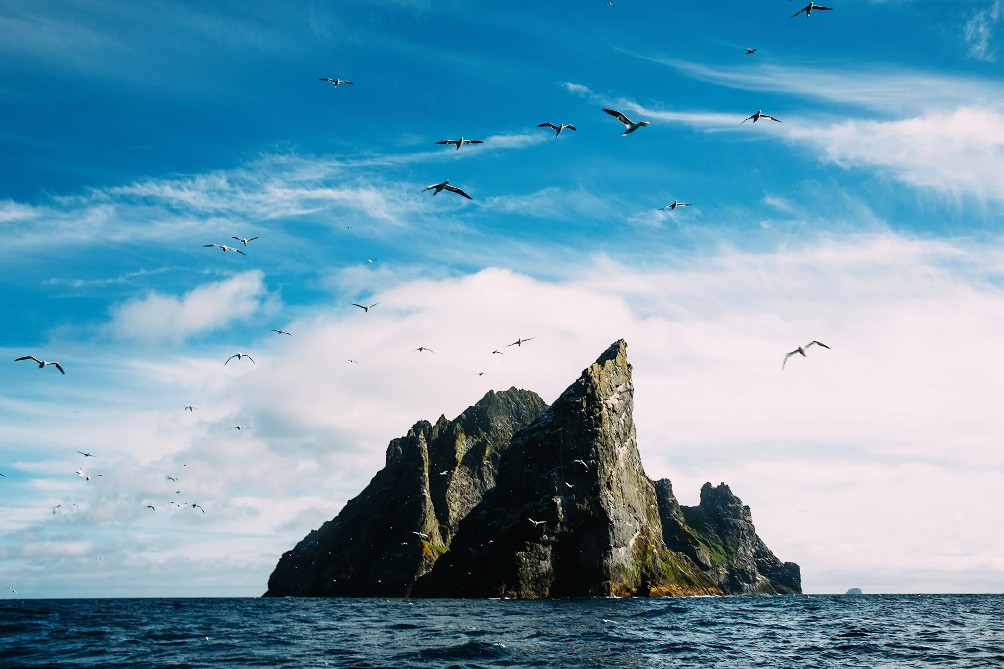 A rocky island rising from the ocean with many seabirds flying in a blue sky with clouds.