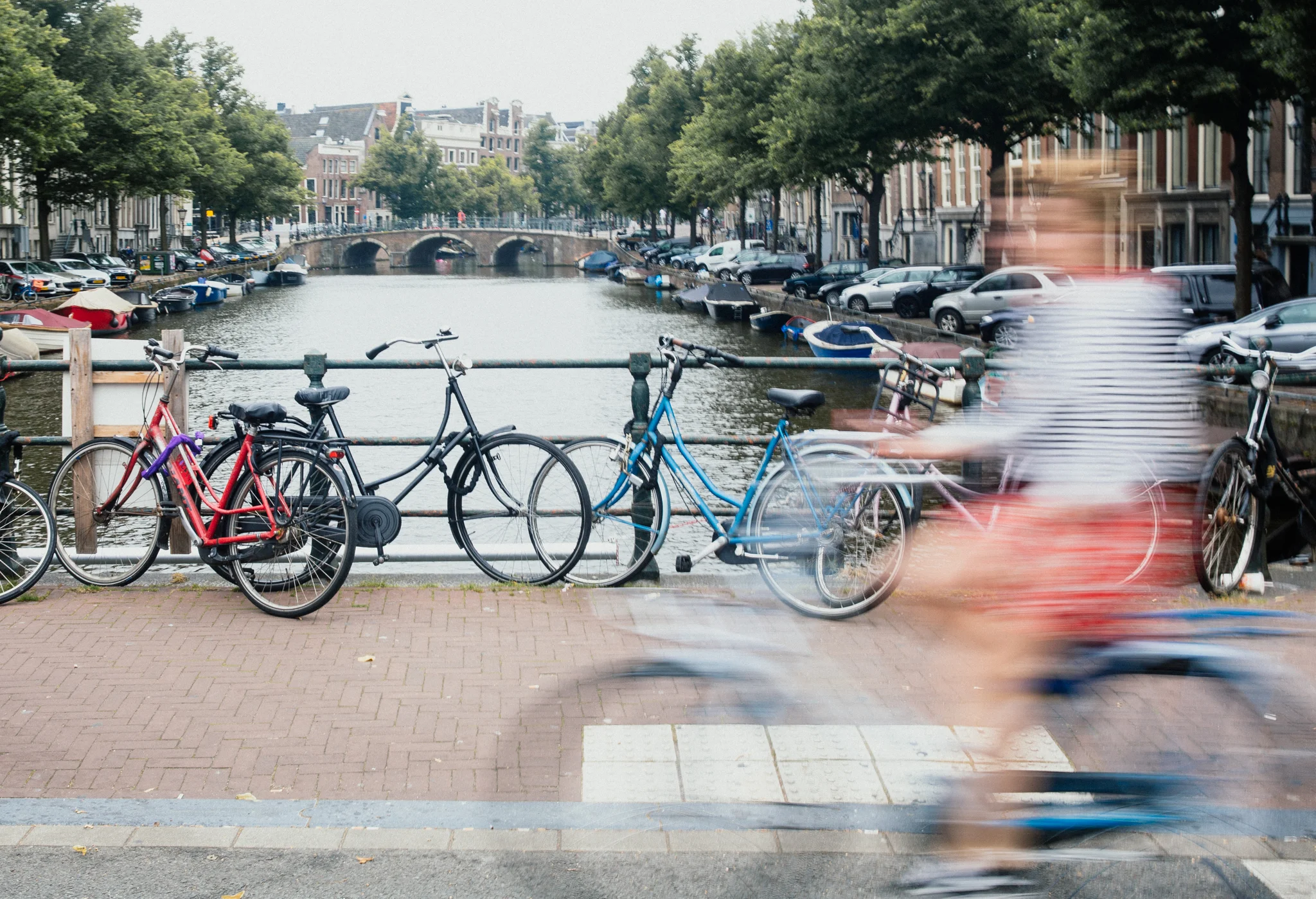 Bicycles parked by a canal with buildings, trees, boats, and a bridge in the background, and a blurred person riding a bike in the foreground.